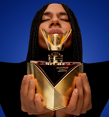 A pair of hands holds a luxurious bottle of Johnnie Walker Vault, showcasing its intricate gold and black design against a vibrant blue background.