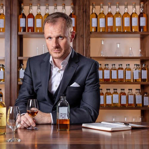 A portrait of Dr. Craig Wilson, dressed in a suit, holding a whisky glass filled with amber liquid. 