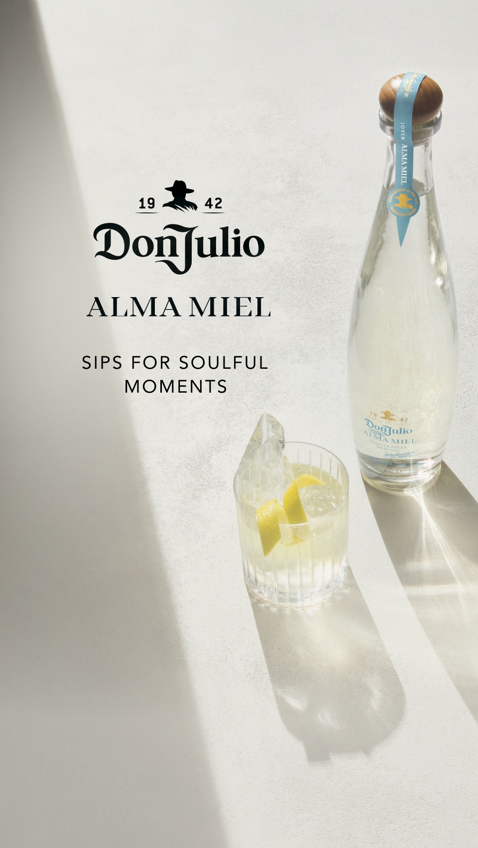 The image features a bottle of Don Julio Alma Miel tequila stands upright on a textured light grey surface.