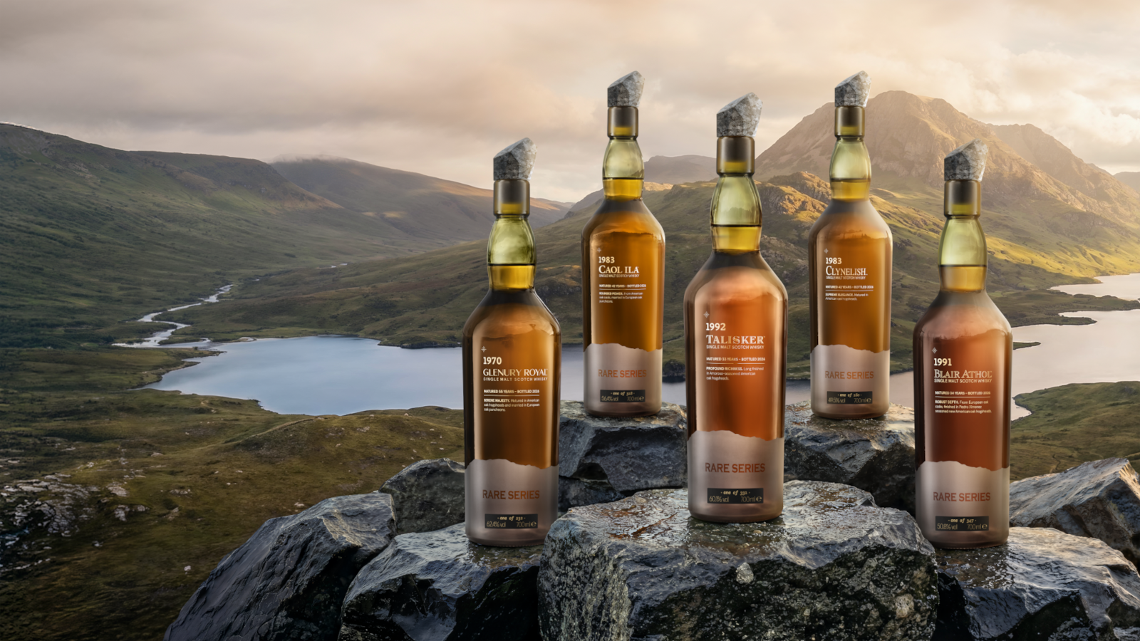 Five whisky bottles on rocky hilltop overlooking lake and mountains at sunset.