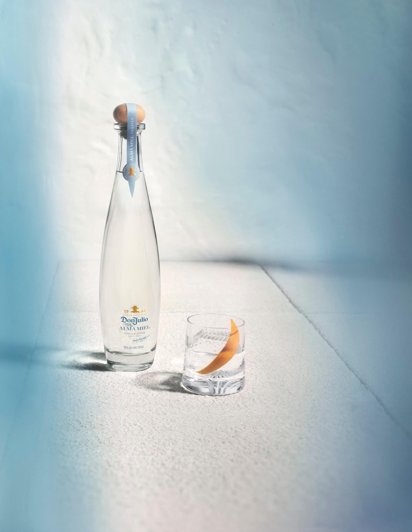 A bottle of Don Julio Alma Miel tequila stands upright on a textured, light-gray surface, with a subtle blue hue cast across the scene.
