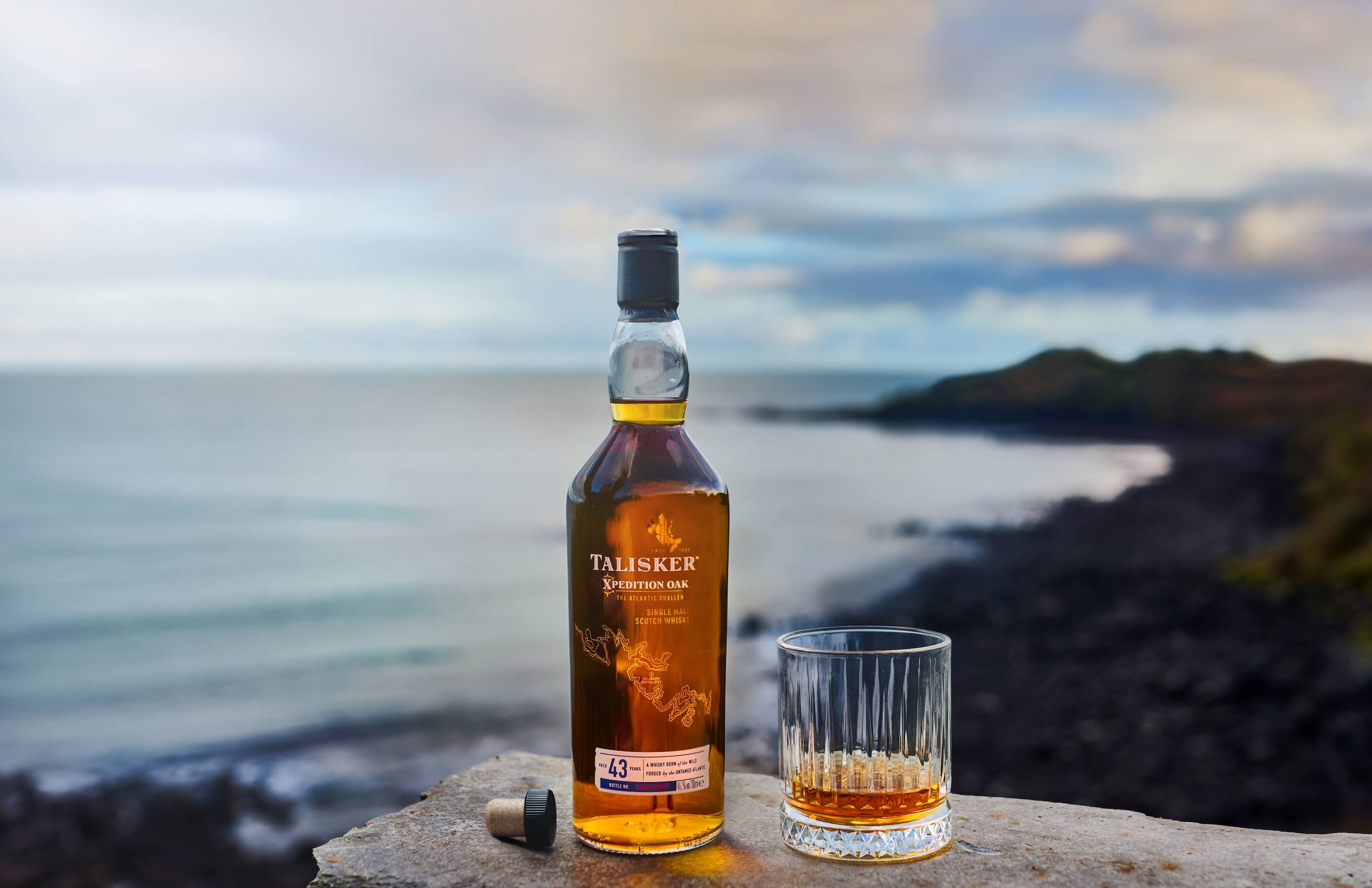 A bottle of Talisker Xpedition Oak 43-Year-Old Single Malt Scotch Whisky is placed on a rocky surface by the sea.
