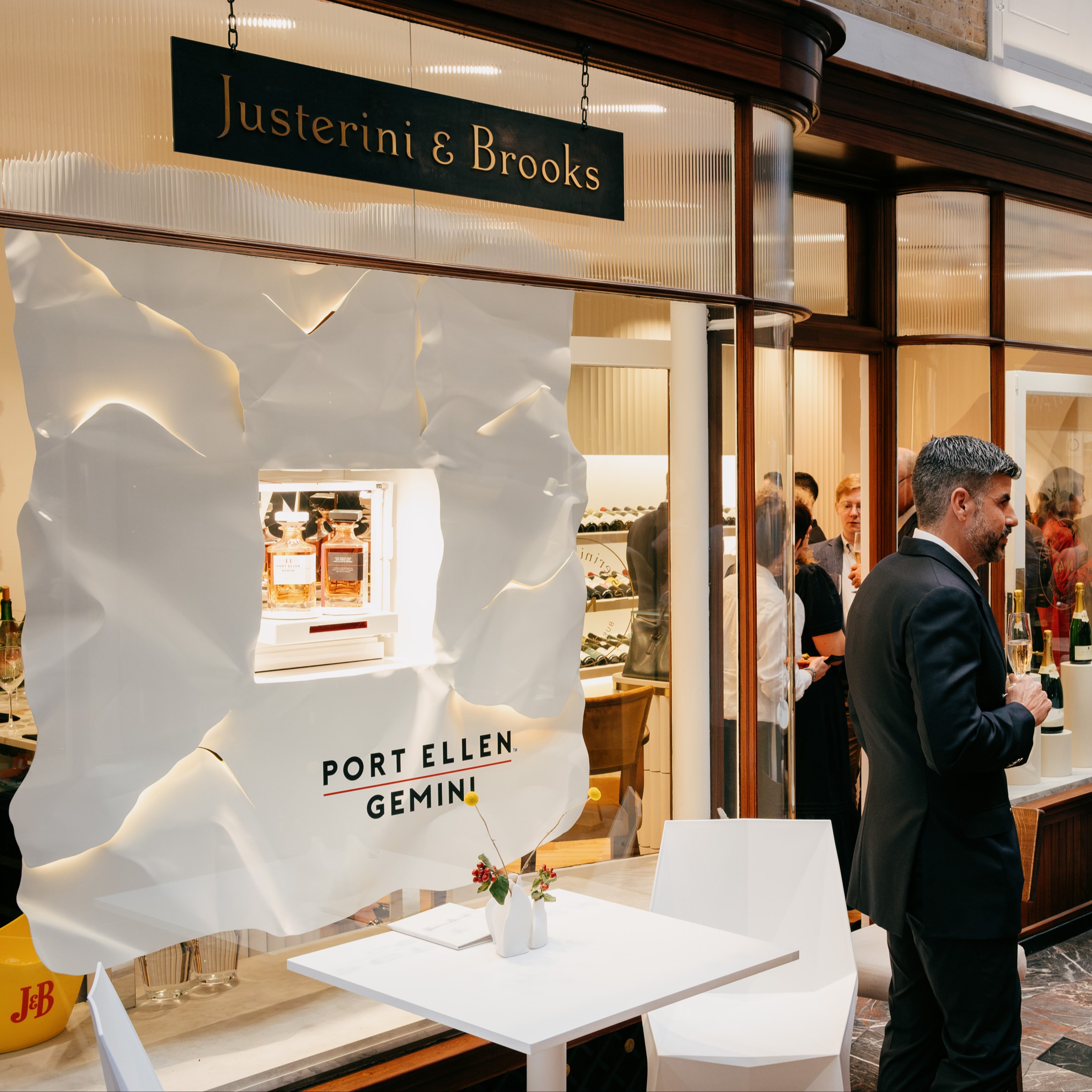 Justerini & Brooks storefront with Port Ellen Gemini display, bottles visible inside. White table and vase in foreground; people gathered inside.
