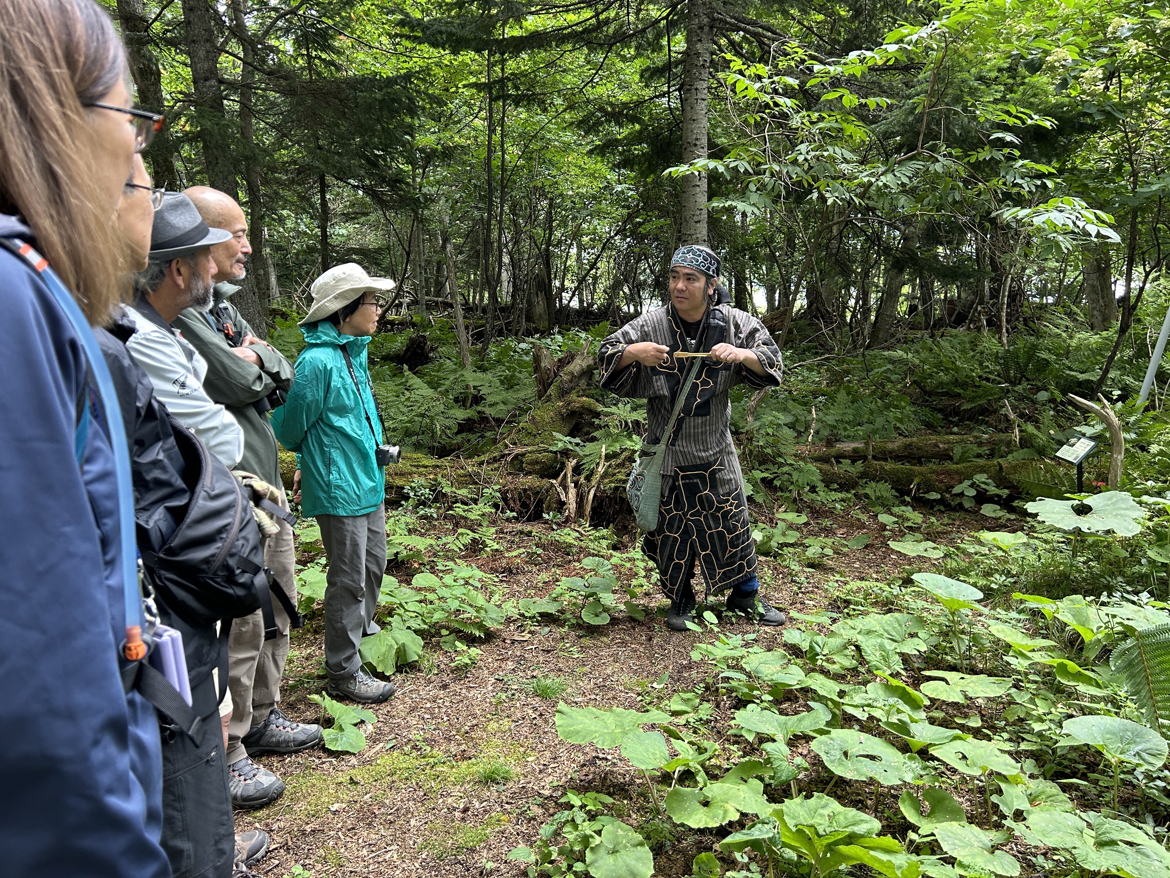 A man dressed in the garb of the Ainu, Hokkaido's indigenous people, speaks to a group of guests on a forest path. He is holding a mukkuri, an Ainu mouth harp made of wood.