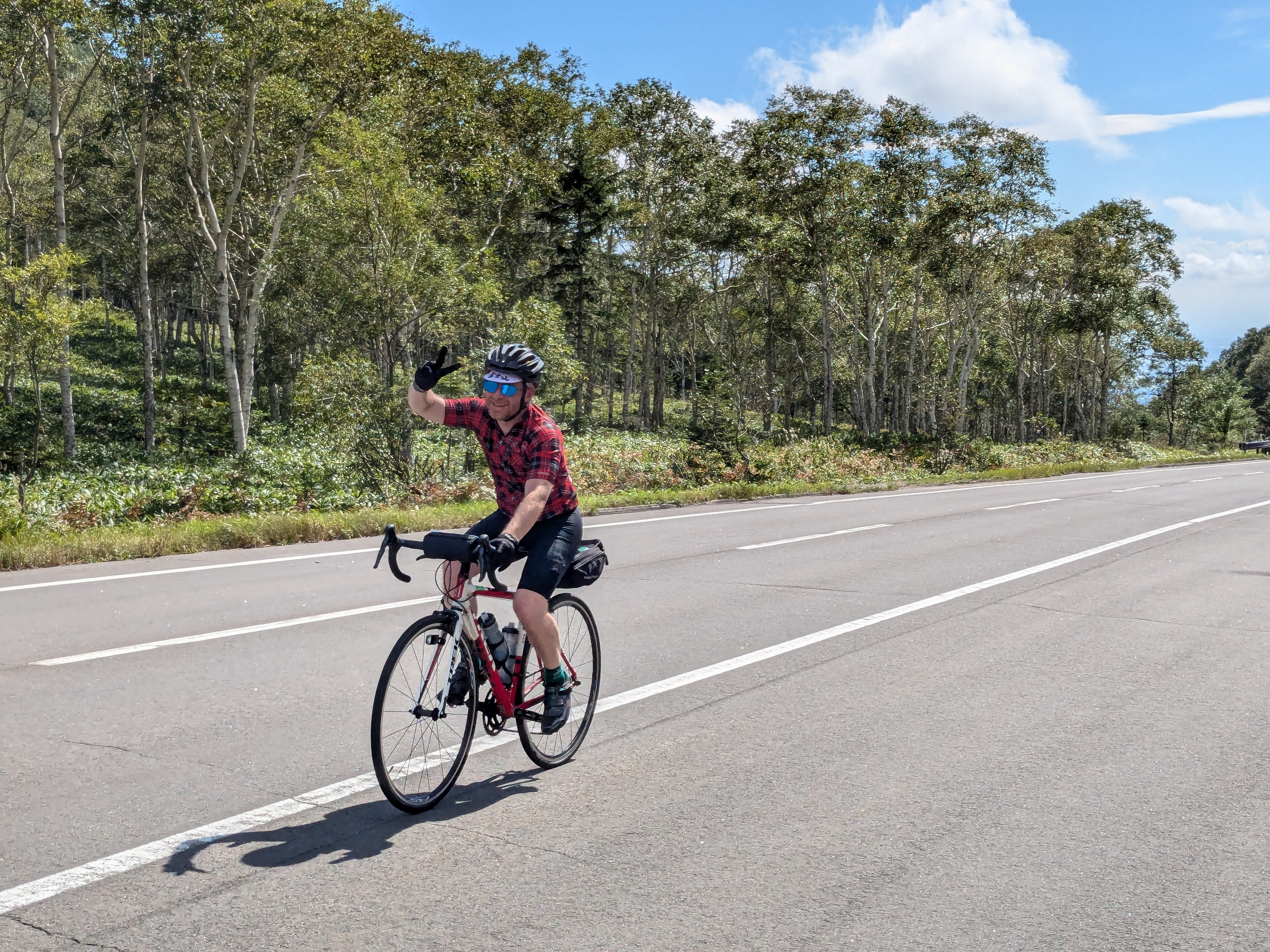 A cyclist on a road raises their hand in the "peace" or "victory" sign (making a V-shape with the index and middle finger).