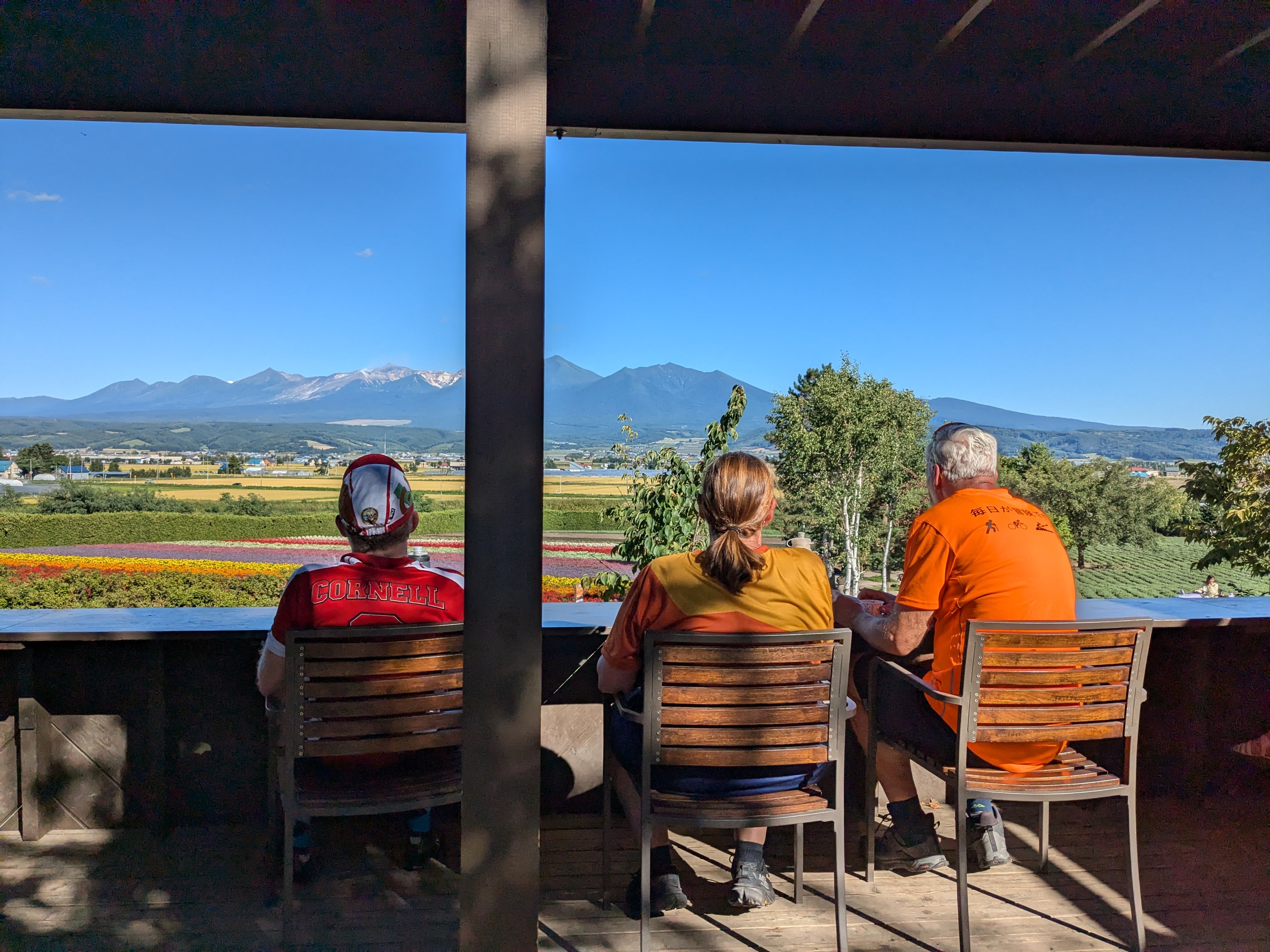 Three cyclists take a break in wooden chairs on a viewing deck, looking out over a field of flowers at the Tokachi Volcanic Group in the distance.