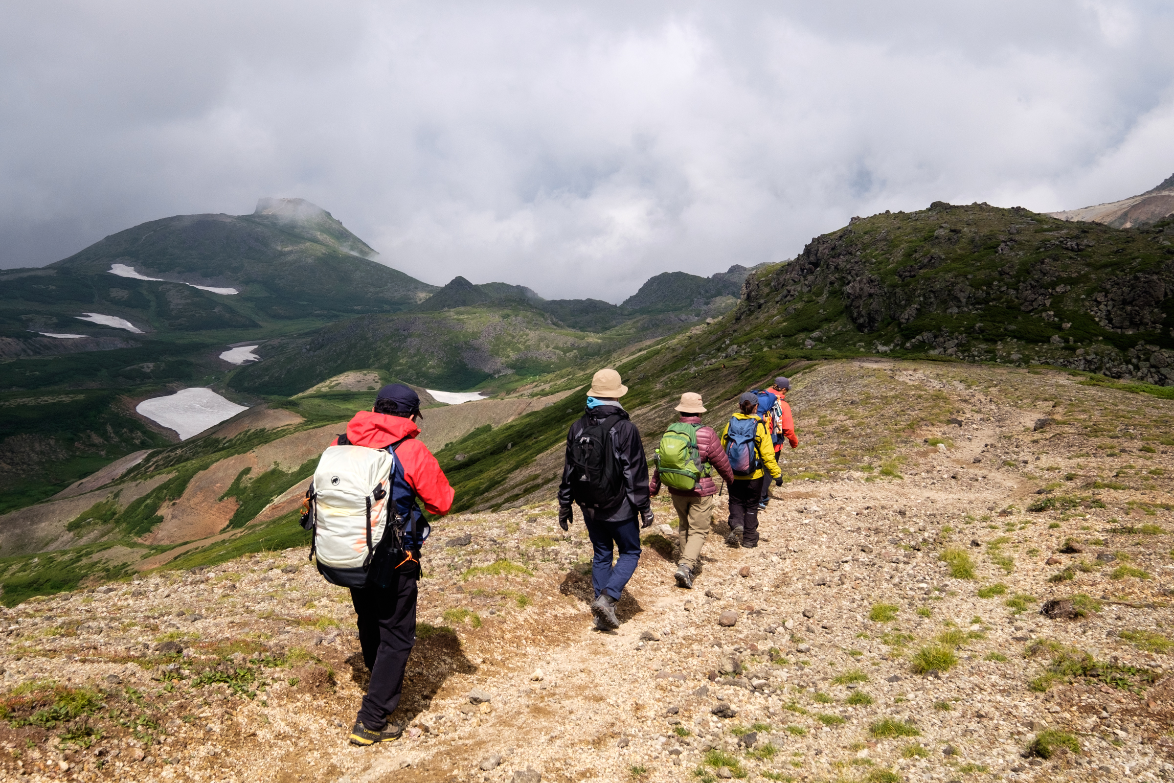 A group of hikers walk along the edge of the Ohachidaira crater in the Daisetsuzan National Park. They are illuminated by afternoon sun while Mt. Kurodake has clouds swirling around it in the background.