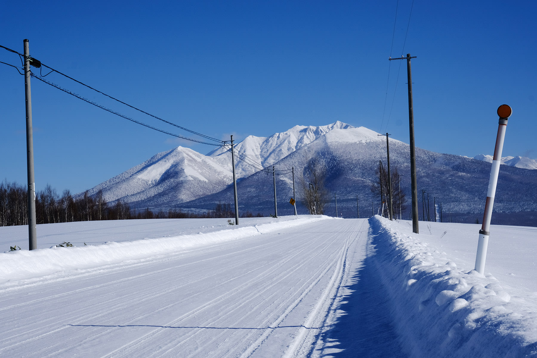 A road is pictured covered in white snow on a clear winters day. A white mountain peak rises above the plains in the background