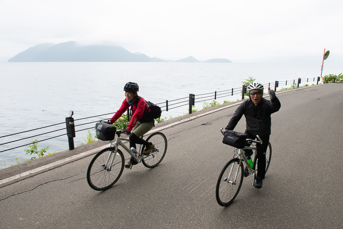 Two cyclists ride along the shore of Lake Toya, with Nakajima in the background