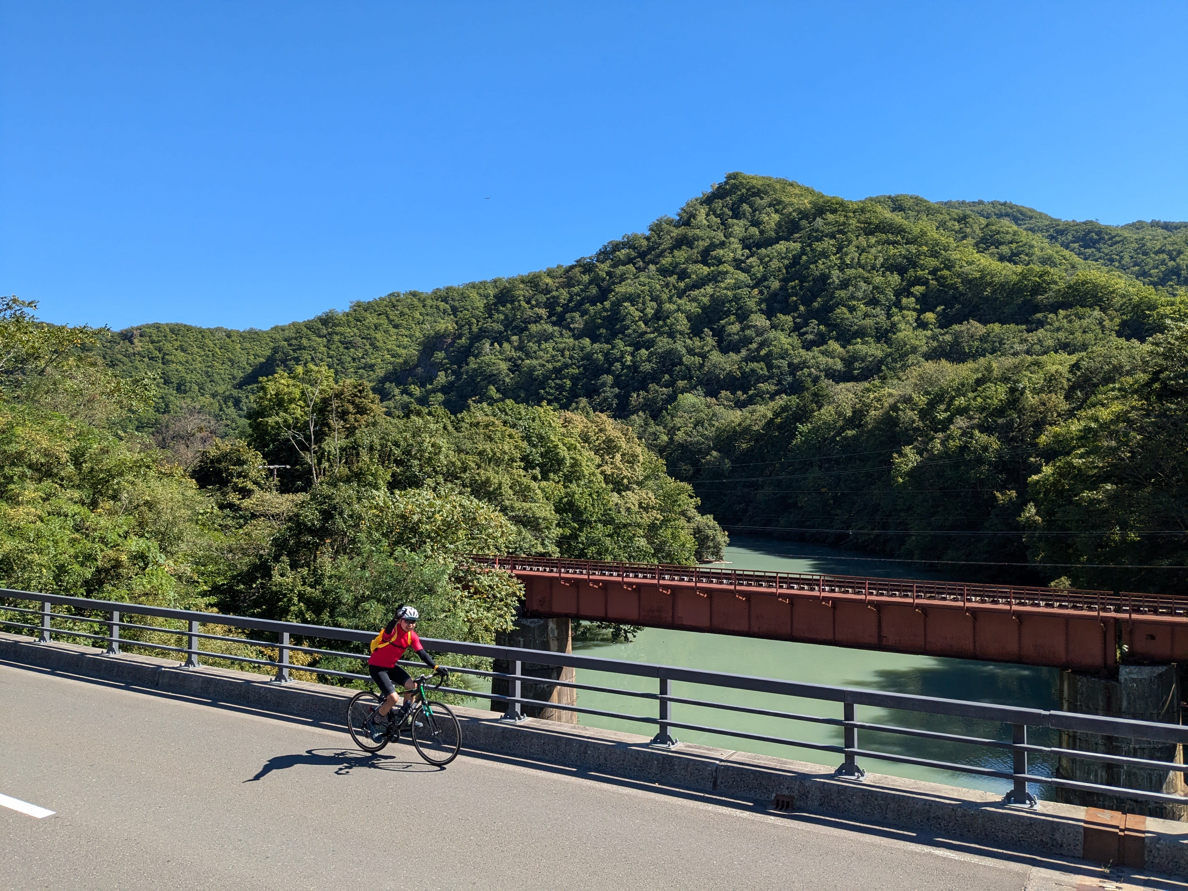 A man on a bike rides over a bridge which spans a river. He is waving to the camera. There is a mountain in the distance behind him and the river. It is a very sunny day.