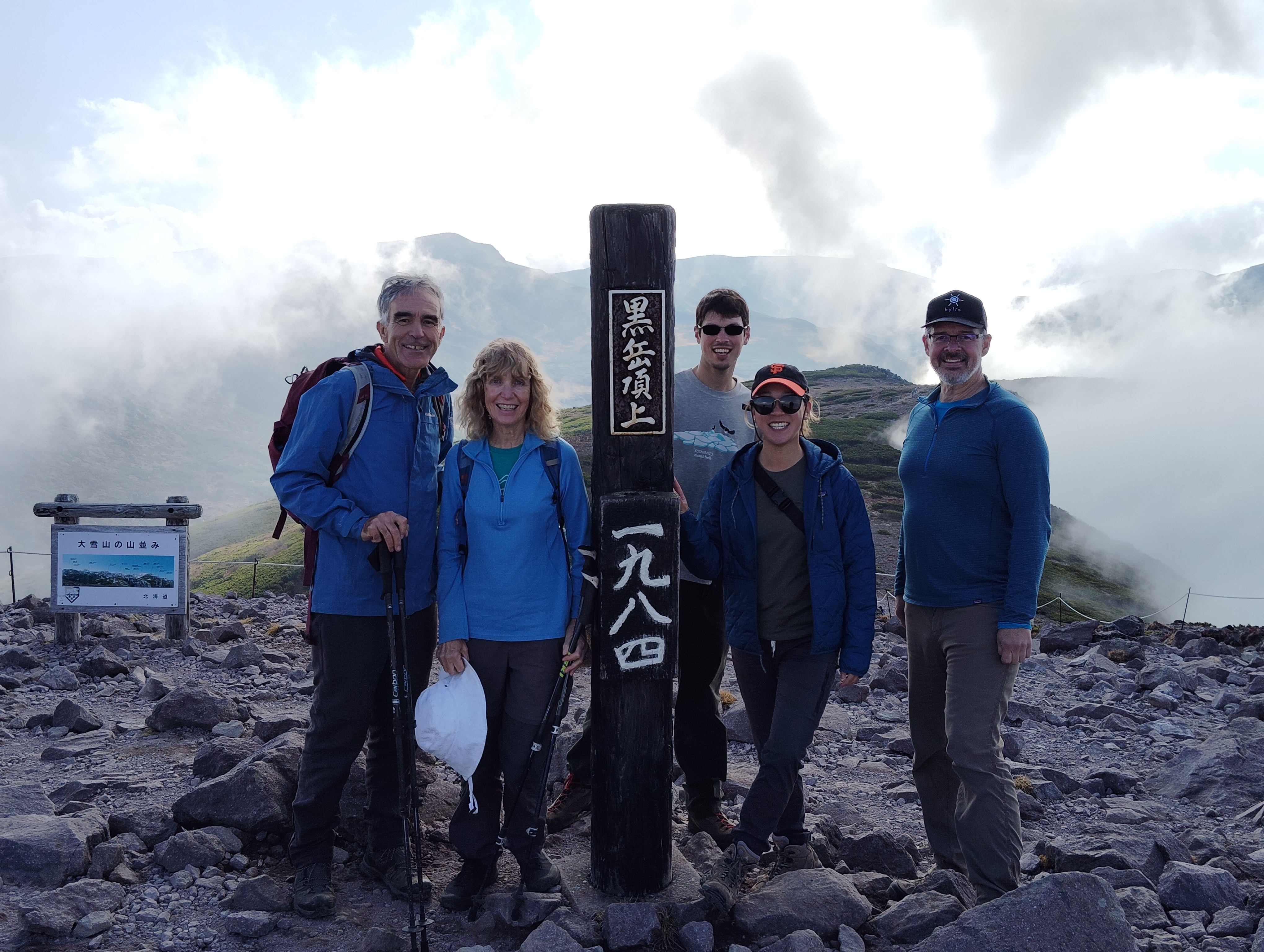 A group of hikers surround a pillar at the summit of Mt. Kurodake in Hokkaido. The pillar reads "Mt. Kurodake Summit, 1980m" in Japanese. The top of the mountain is quite foggy.