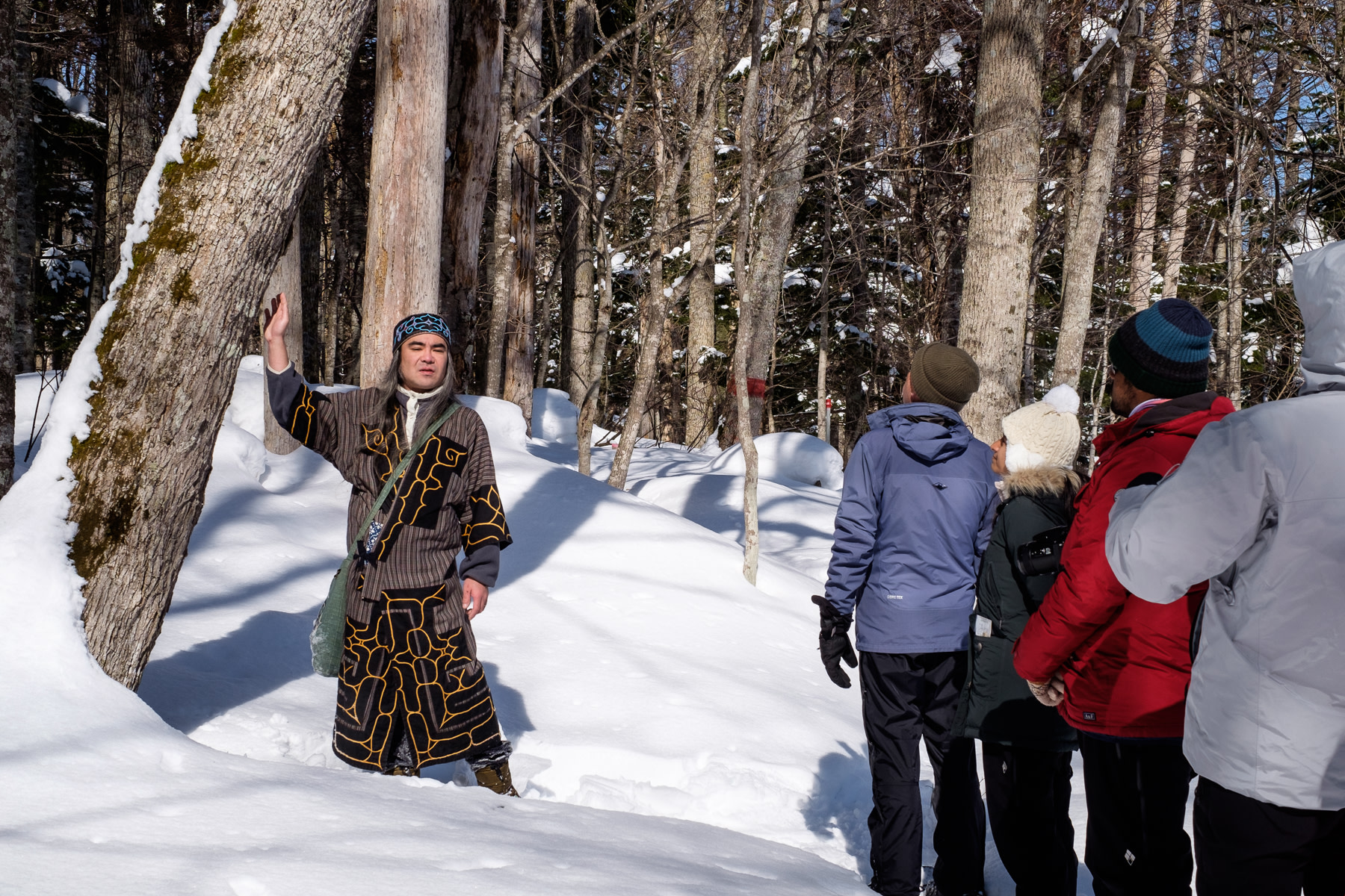 An Ainu guide wearing Ainu clothing stands next to a tree while explaining the uses for the wood to a group of guests in the Akan–Mashu National Park