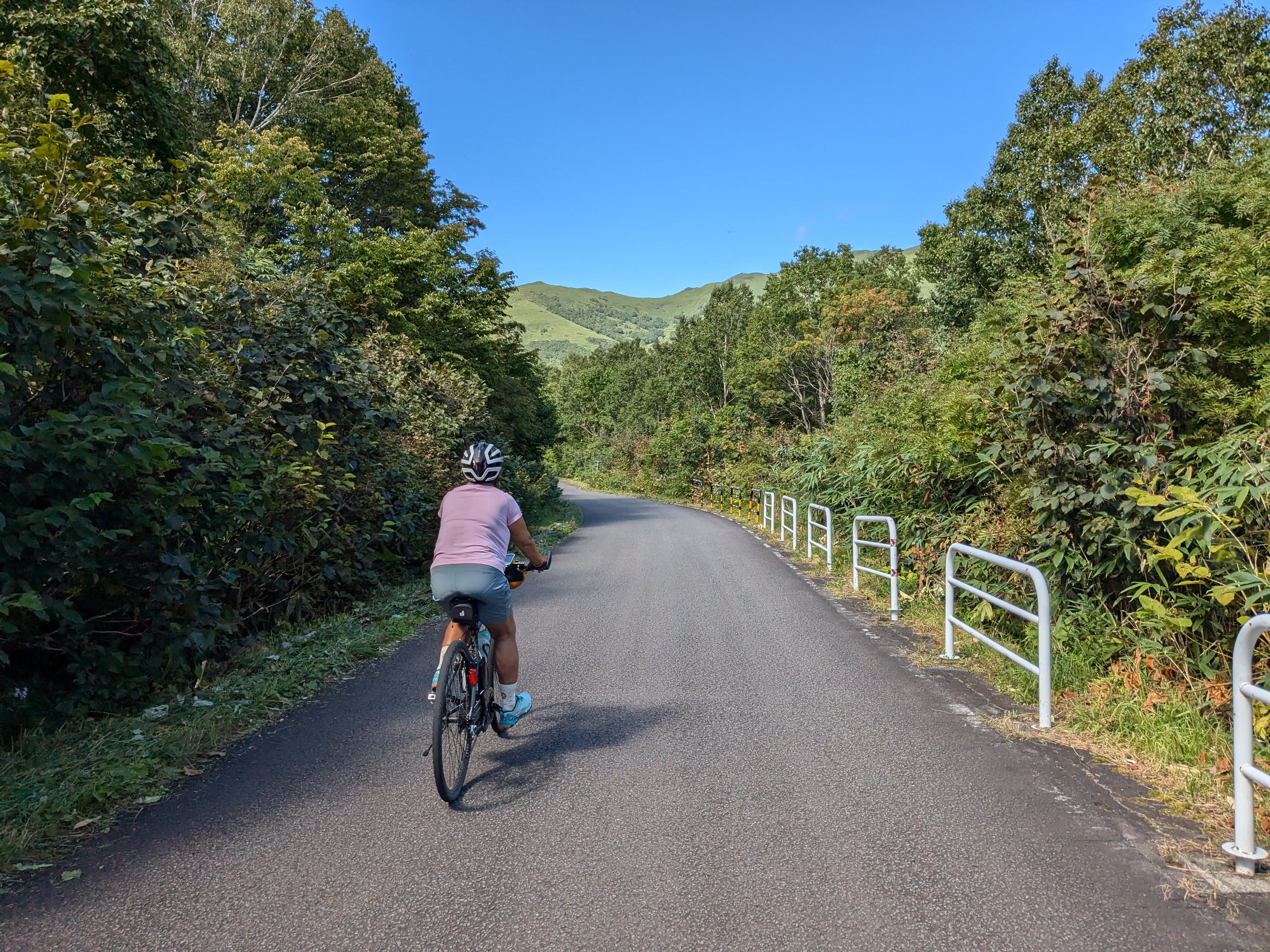 A woman cycles on a road through a mountain pass. There are the green hills of mountain highlands in the distance. It is a beautifully clear, sunny day.