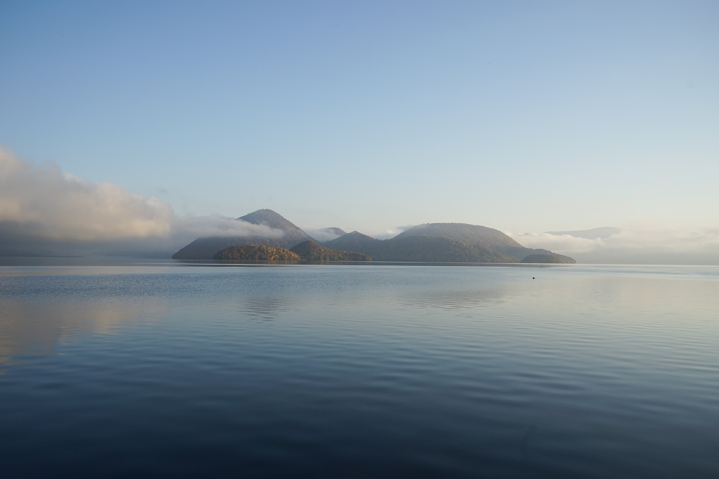 A photo of Nakajima Island in the middle of Lake Toya. It is the early morning and the water is very still, with mist rising over the island.