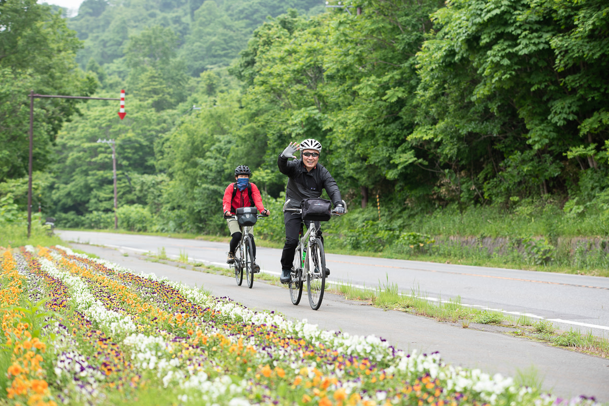 A cyclist waves whilst riding past rows of flowers