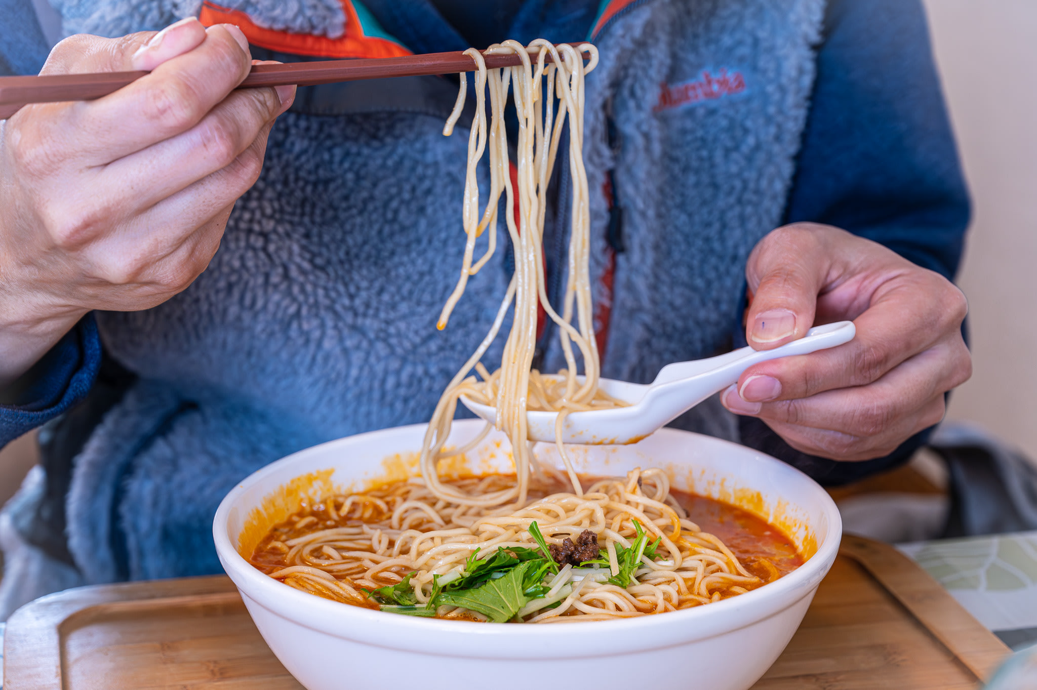 A man's hands use chopsticks and a spoon to lift noodles from a bowl of dandan ramen (spicy Sichuan-style ramen). The broth is bright red and looks very spicy.