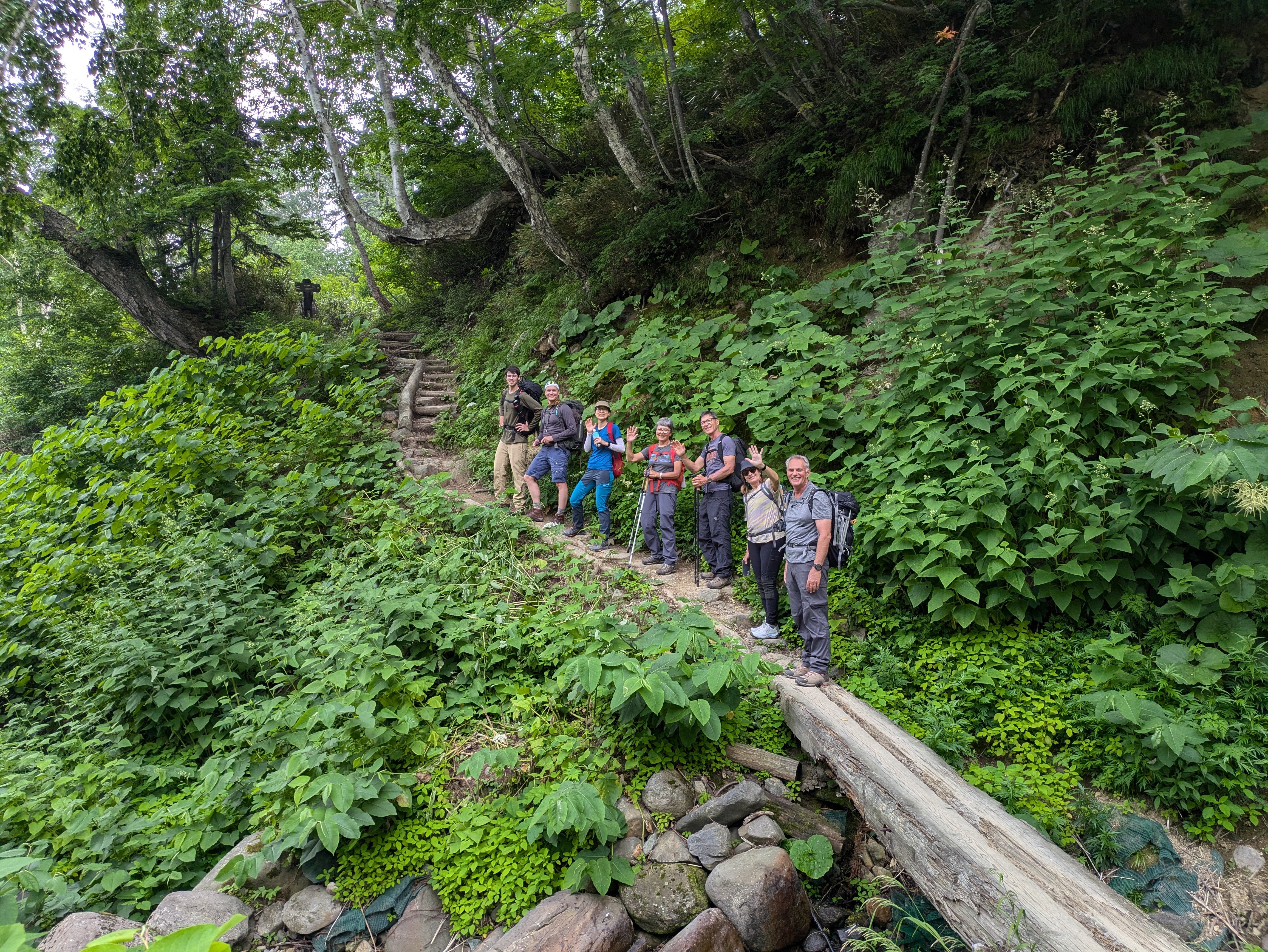 A line of smiling hikers on a mountain trail ahead. They are all facing back to the camera and smiling.