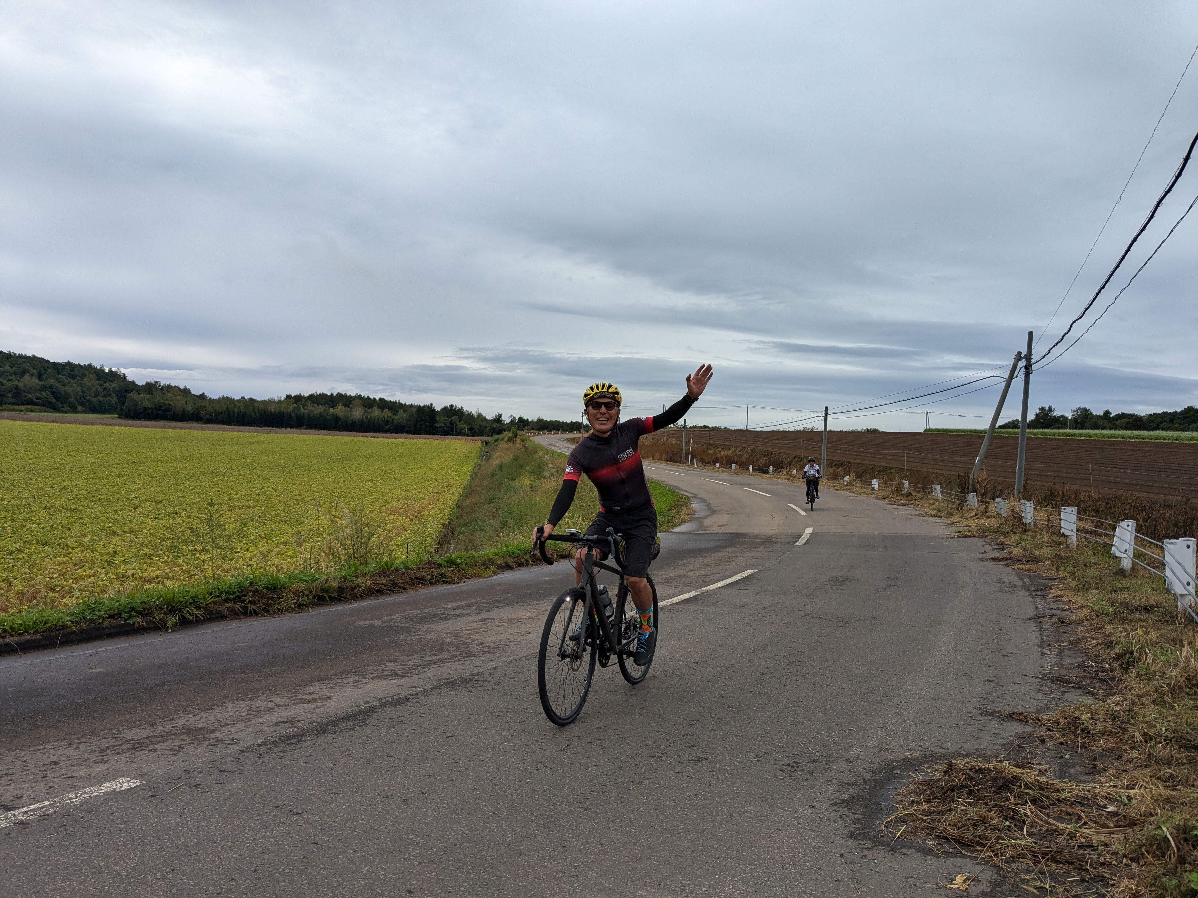 A man waves to the camera as he cycles on a road alongside a soy bean field in southern Hokkaido. It is an overcast and cloudy day.