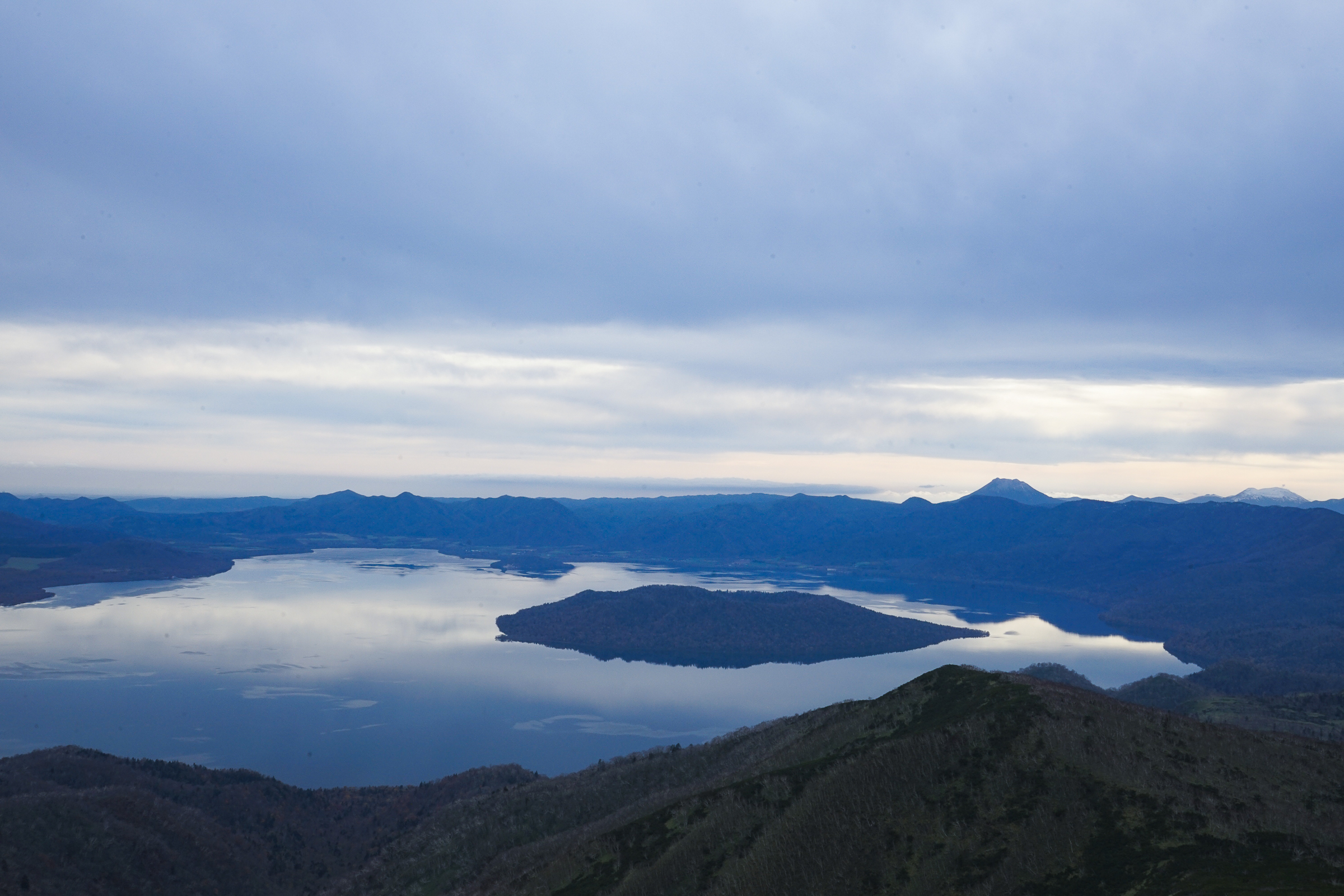 Nakajima island floats in the middle of Lake Kussharo on a calm day. Viewed from high on Mt Mokoto, the lake seems like a mirror, reflecting the other peaks of the Akan–Mashu National Park