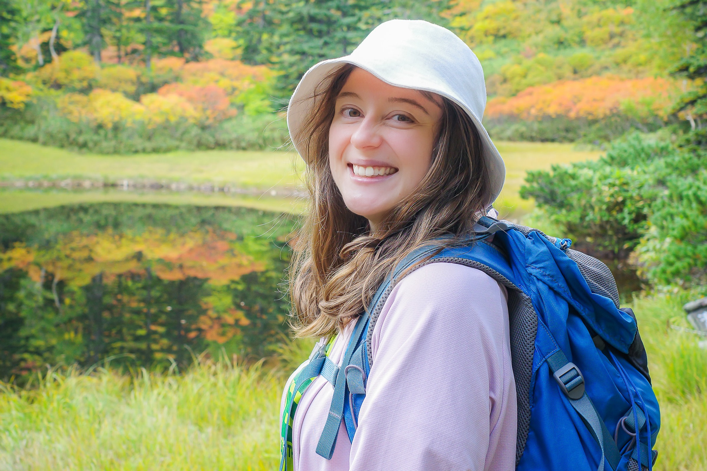A female hiker wearing a cute white hat beams into the camera. In the background the first autumn colours in the Daisetsuzan are reflected in a still pond at Kogen Onsen.