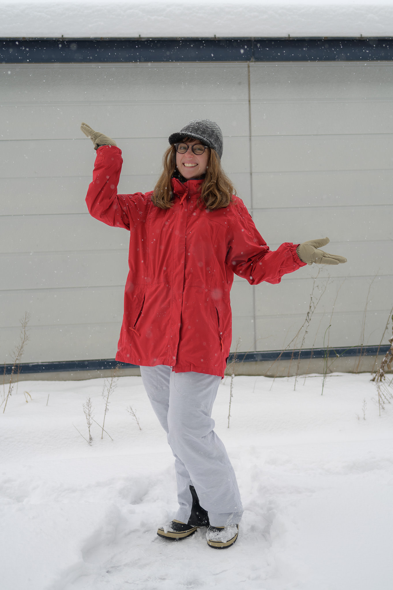 A girl wearing glasses poses in the snow in a waterproof outdoor jacket, a beanie with a cap, ski gloves and ski pants.