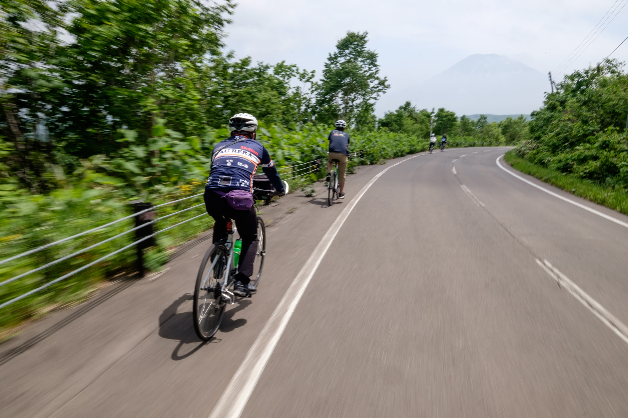 Cyclists descend a hill at speed with Mount Yotei in the background