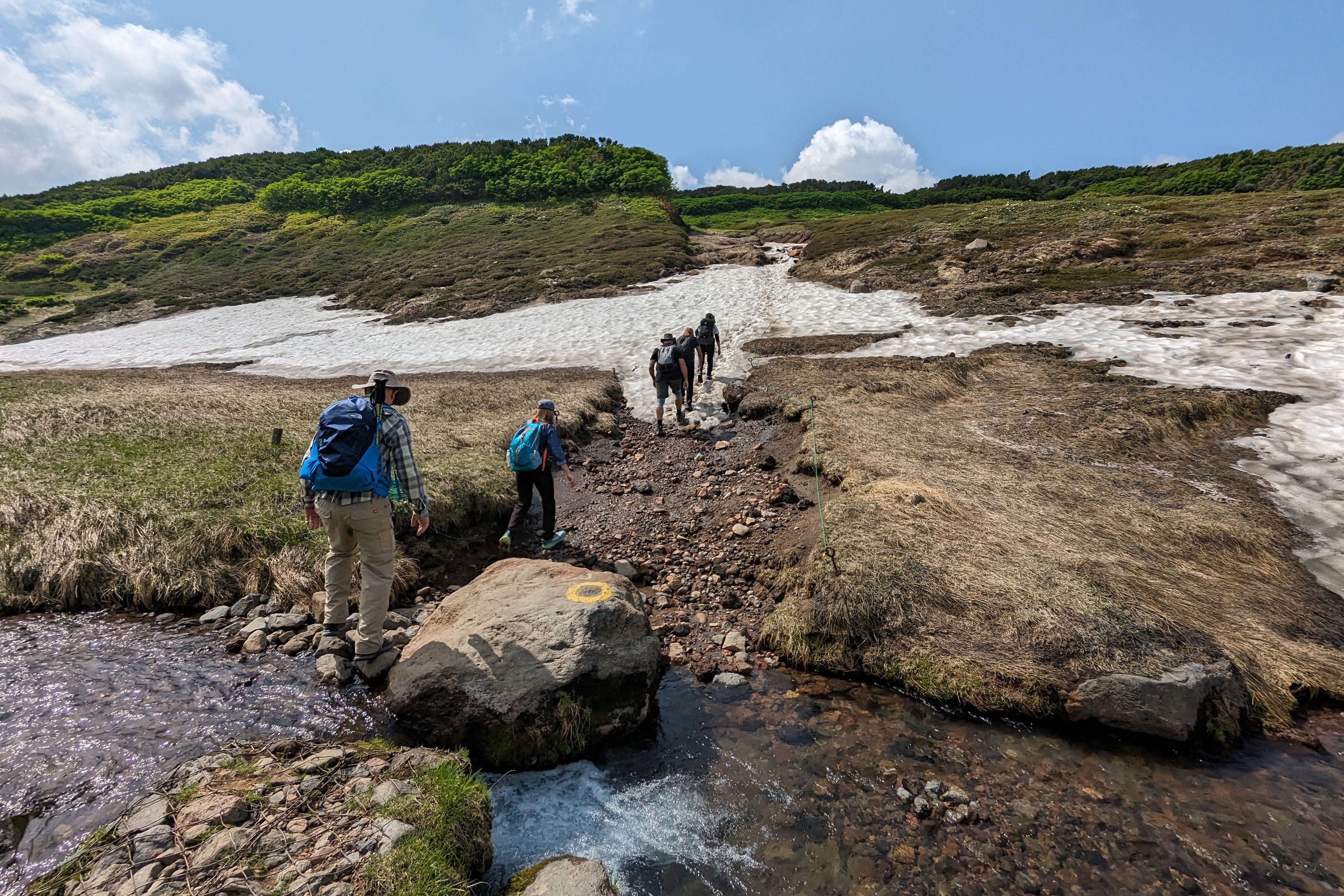 A group of hikers traverse difficult terrain, including a small stream, loose rocks and a snowfield.