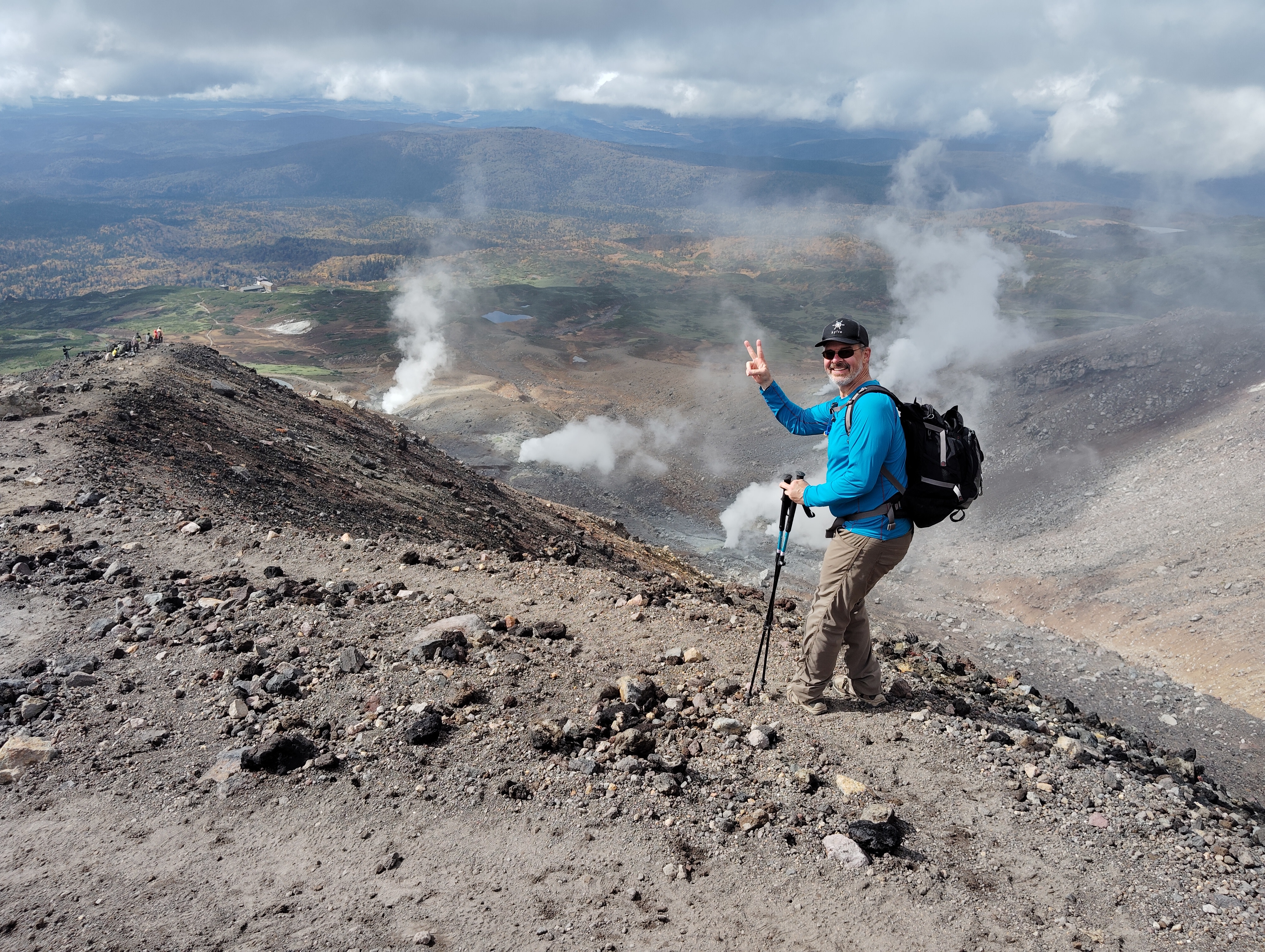 A man gives a "peace" or "victory" sign (forefinger and middle finger raised in a "V" shape) to the camera on a rocky mountain trail. Behind him, volcanic fumaroles are visible in the distance, sending up columns of steam. 