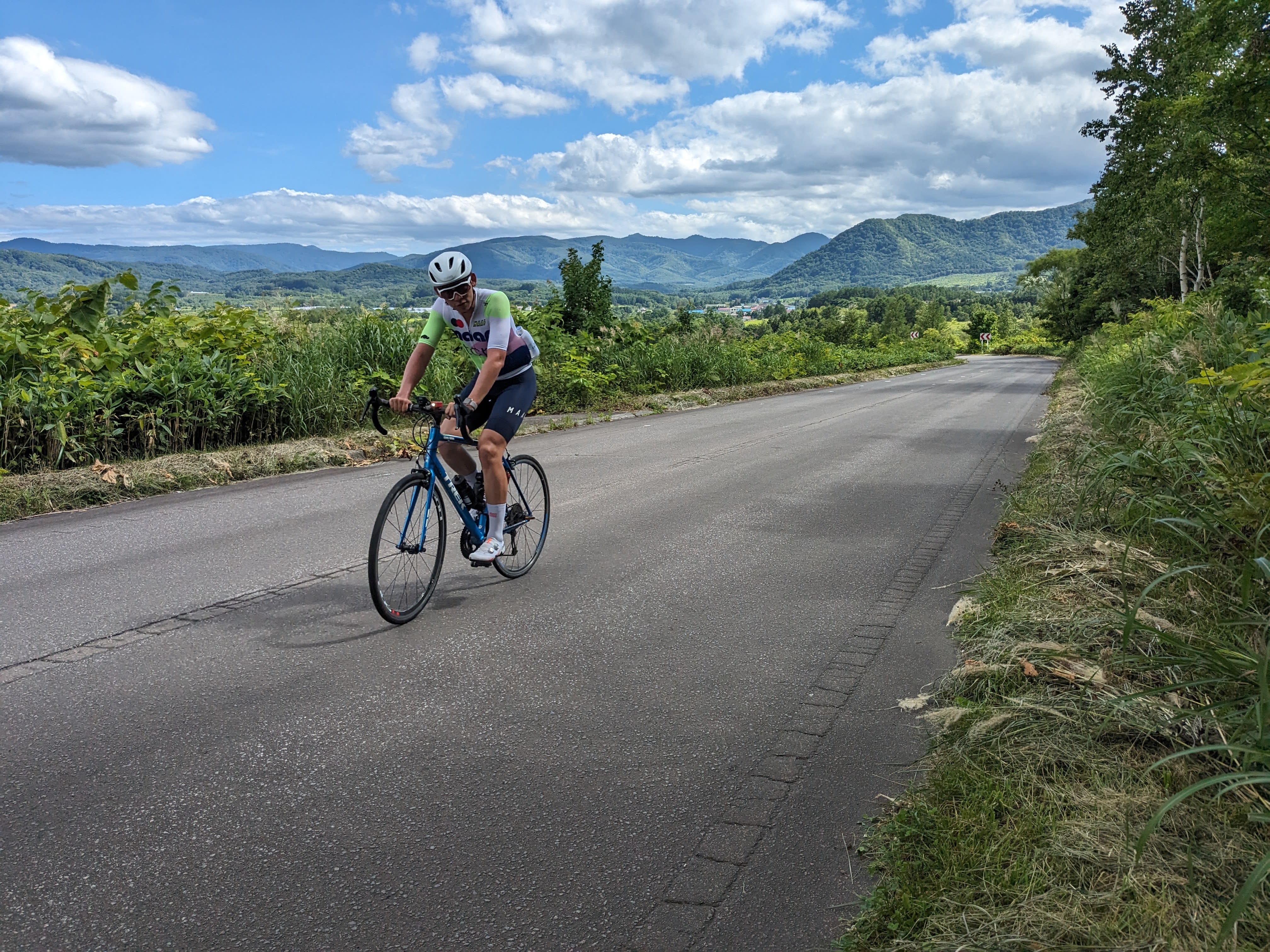 A cyclist in bright cycling gear rides down a country road, with blue skies and white clouds trailing away behind him.