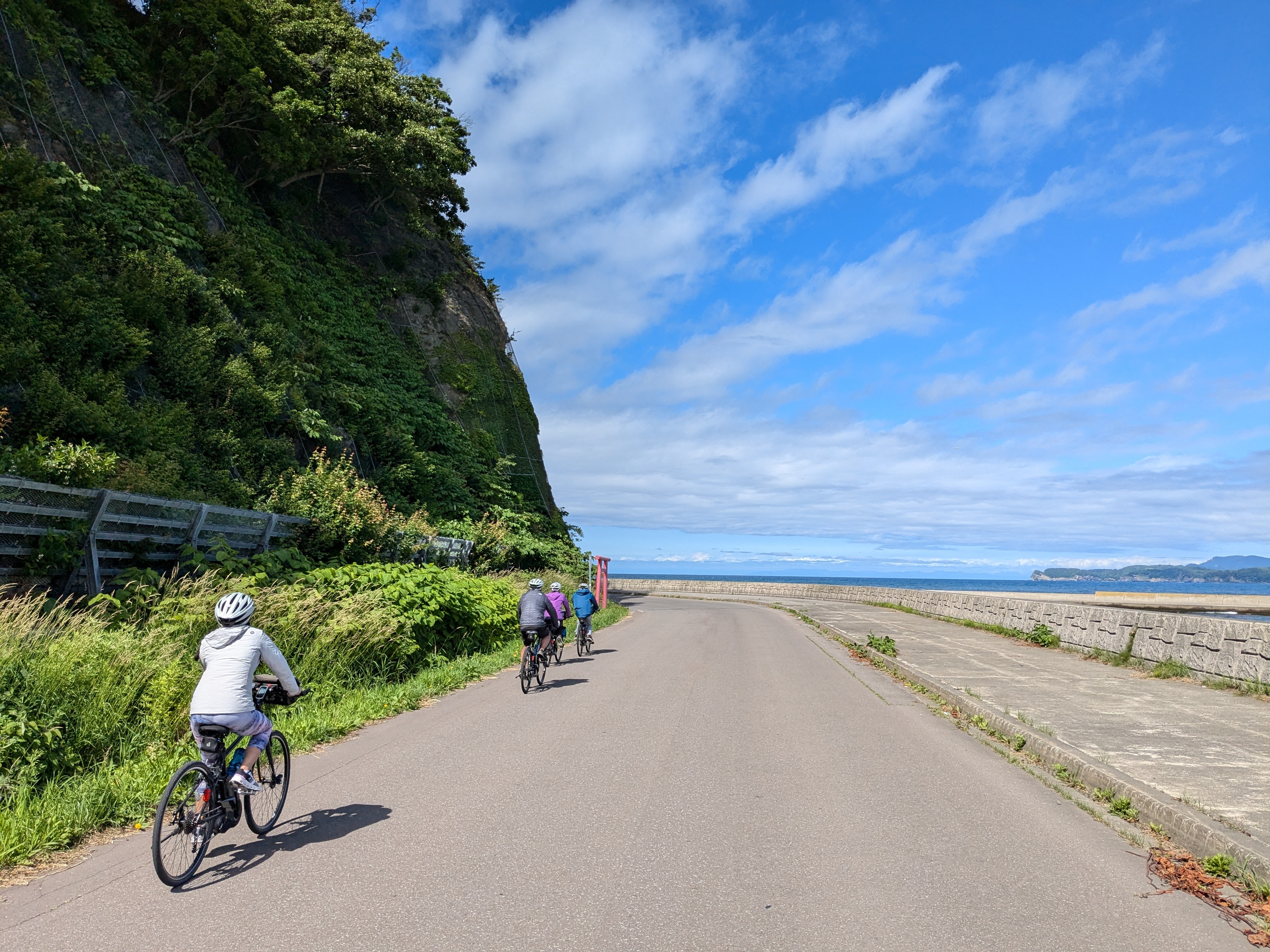 A group of four cyclists rides along a road, approaching the coast and the Sea of Japan beyond. It is a very sunny day.