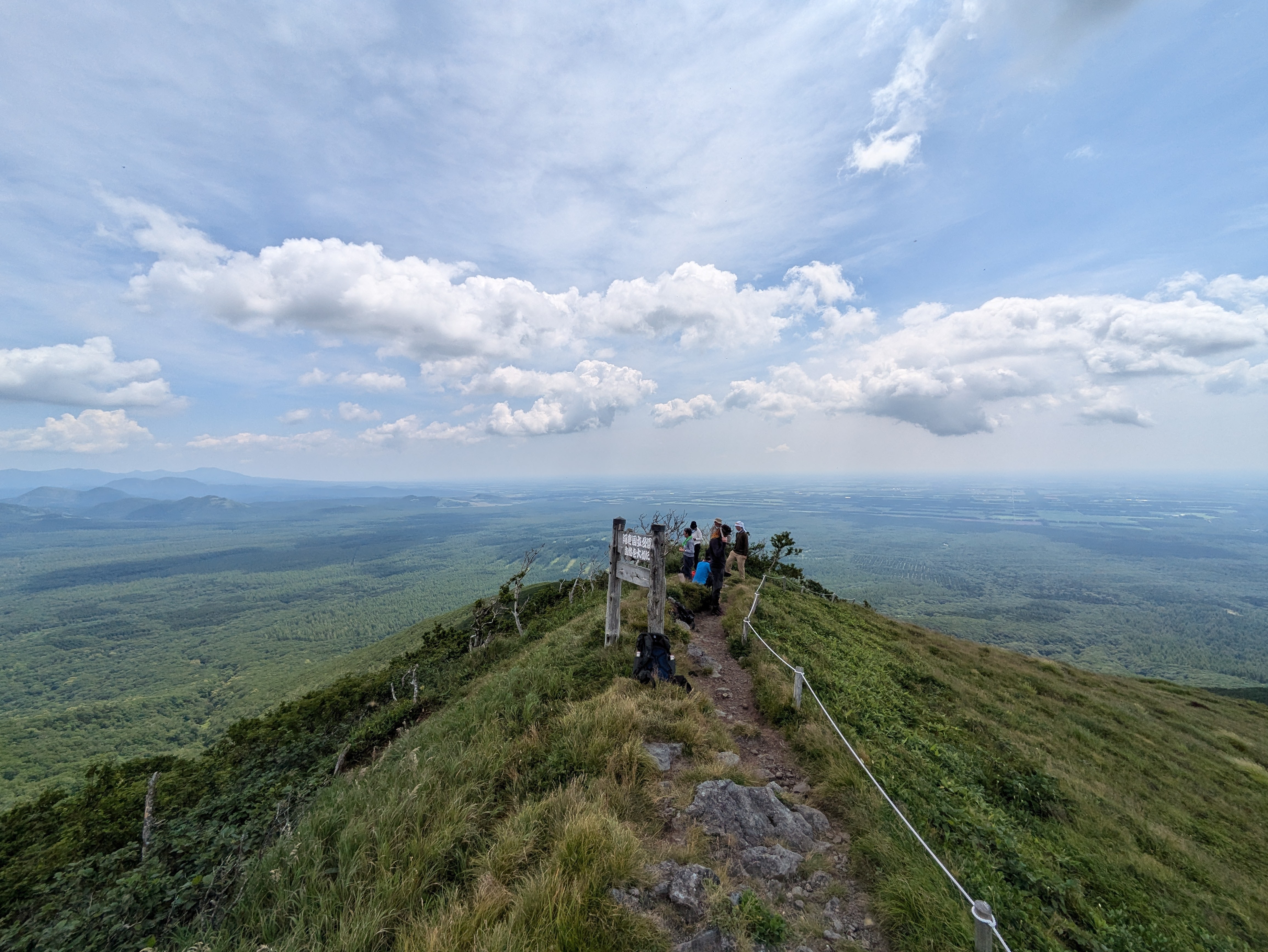 A wide-angle view of hikers at the summit of Mt. Nishibetsu on a very clear day. They all stand on the narrow peak, a flat expanse stretching far away below them.