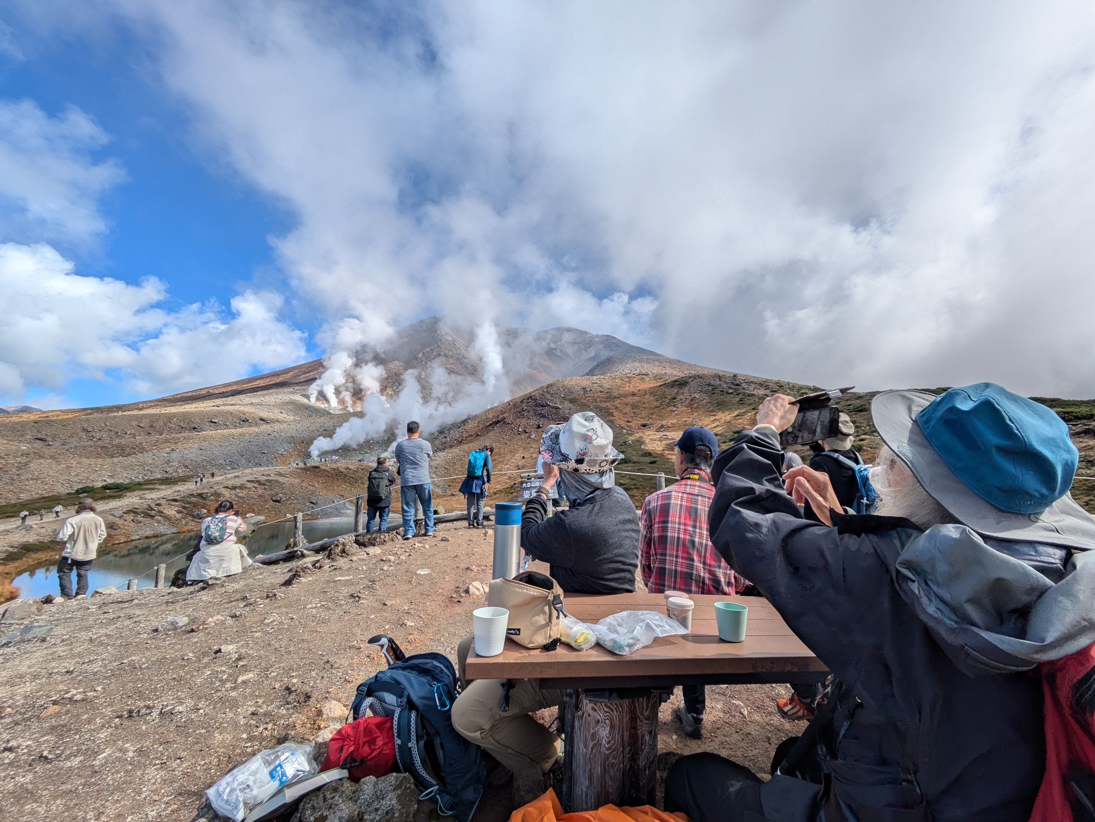 Two ladies sit at a picnic table in view of Sugatami Pond on Mt. Asahidake, Hokkaido. There are many other people there looking at the pond as well. The two ladies are taking a photo of the pond and steaming volcanic vents behind it. Their lunch, a picnic, is on the table in front of them.