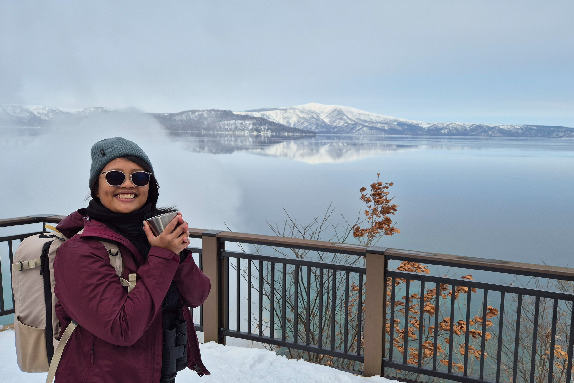 A female tourist enjoying the winter scenery at Oyakotsu Jigoku observation deck on Wakoto Peninsula, overlooking the calm surface of Lake Kussharo. Snow-capped Mount Mokoto rises majestically in the background. The visitor, bundled in winter attire, warms herself with a hot beverage while smiling at the camera.