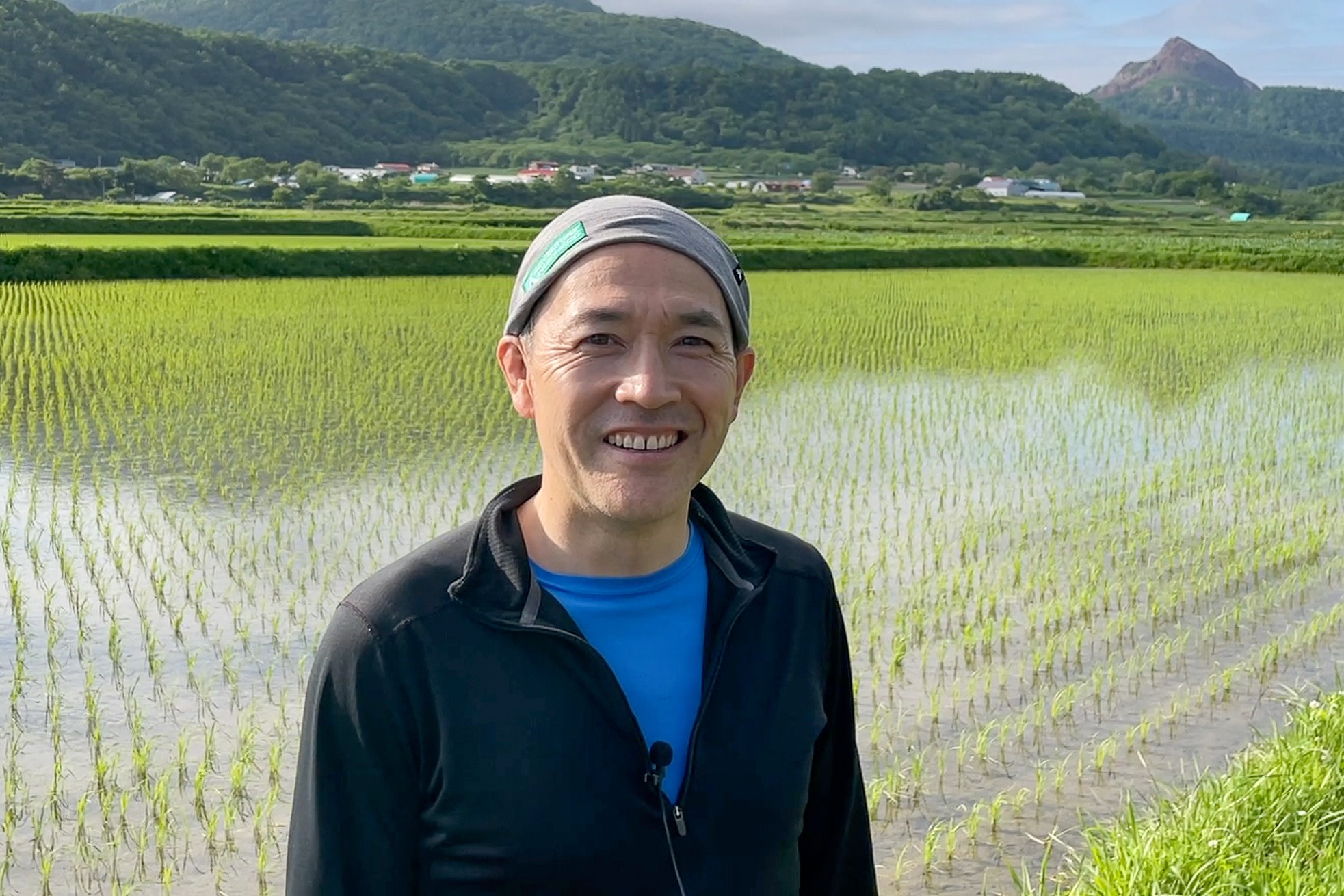 A cycling guide smiles to the camera while standing amongst flooded rich fields. In the distance is the jagged volcanic peak of Mt. Showashinzan.