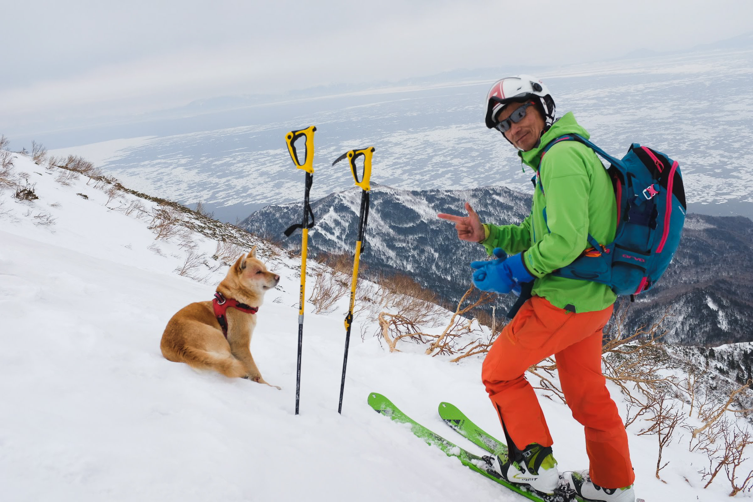 Adventure Hokkaido guide Gen poses high on a mountain while skiing in Shiretoko. He is with his dog, a tea coloured Shiba Inu. In the background, drift ice floats on the surface of the Sea of Okhotsk.