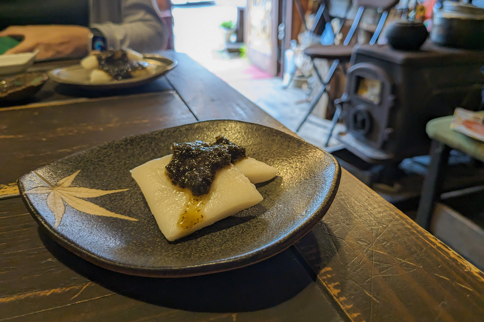 A small "otoshi" dish on a side plate. It is a dish of two small pillows of mochi topped with a dark sauce.