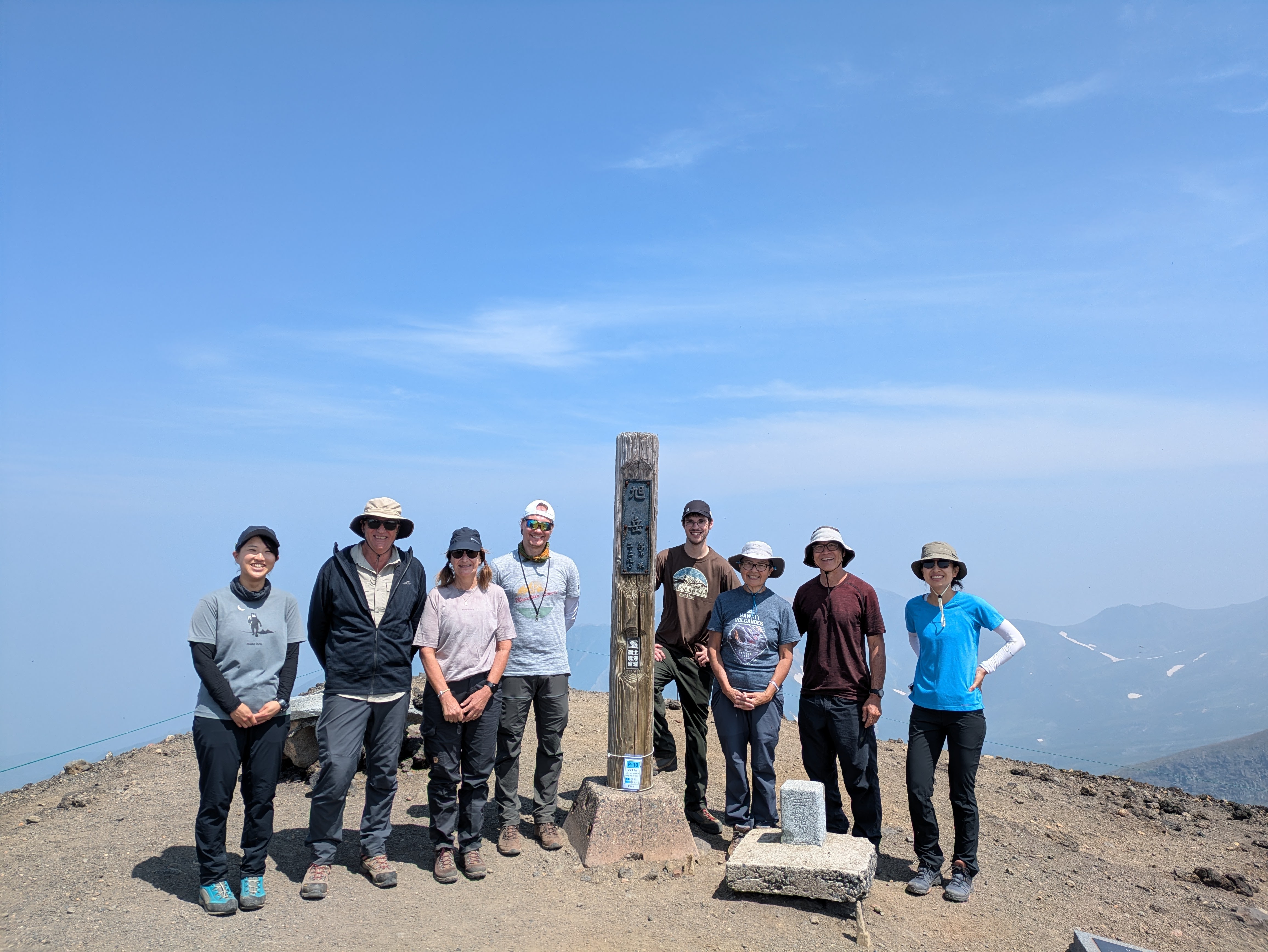 A group of hikers at the summit of a mountain. A pole in Japanese reads "Asahidake, 2290m". They are all smiling at the camera.