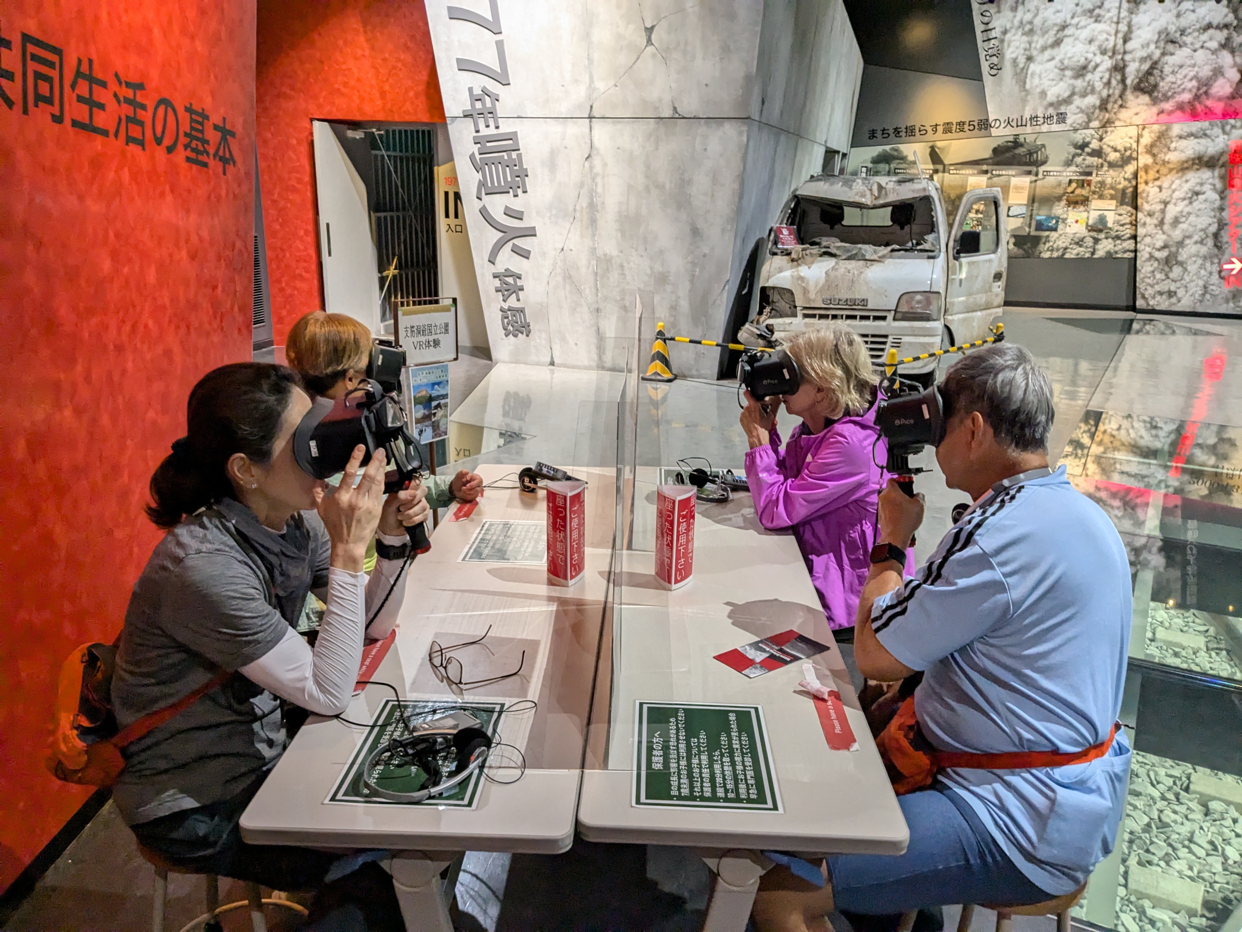 A group of four people at the Lake Toya Volcano Science Museum sit at a table with virtual reality devices strapped on their faces. These machines will allow them to experience aspects of volcanic eruptions and Lake Toya as if they were really there.