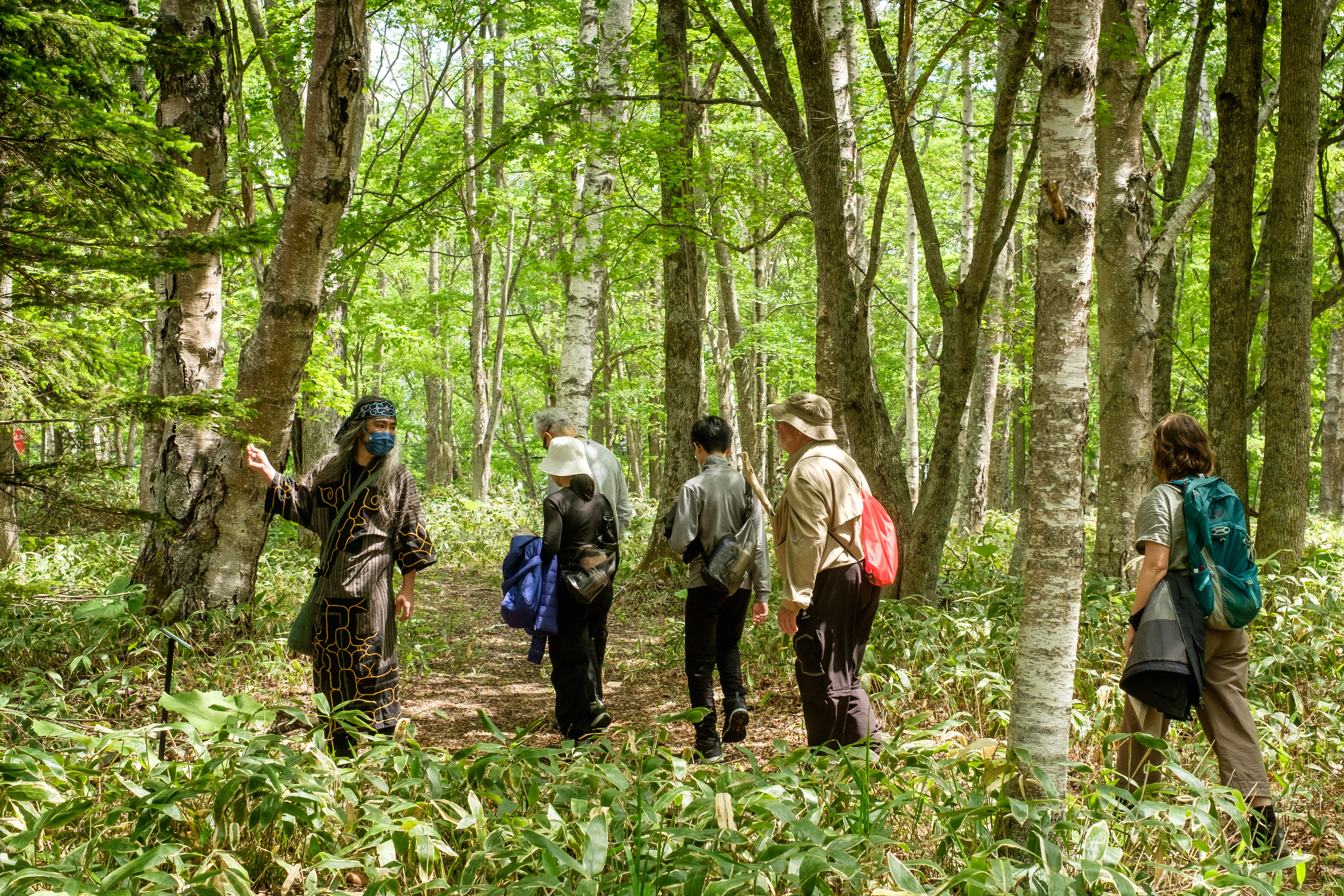 An Ainu guide in traditional garb leads a group of visitors through a forest.