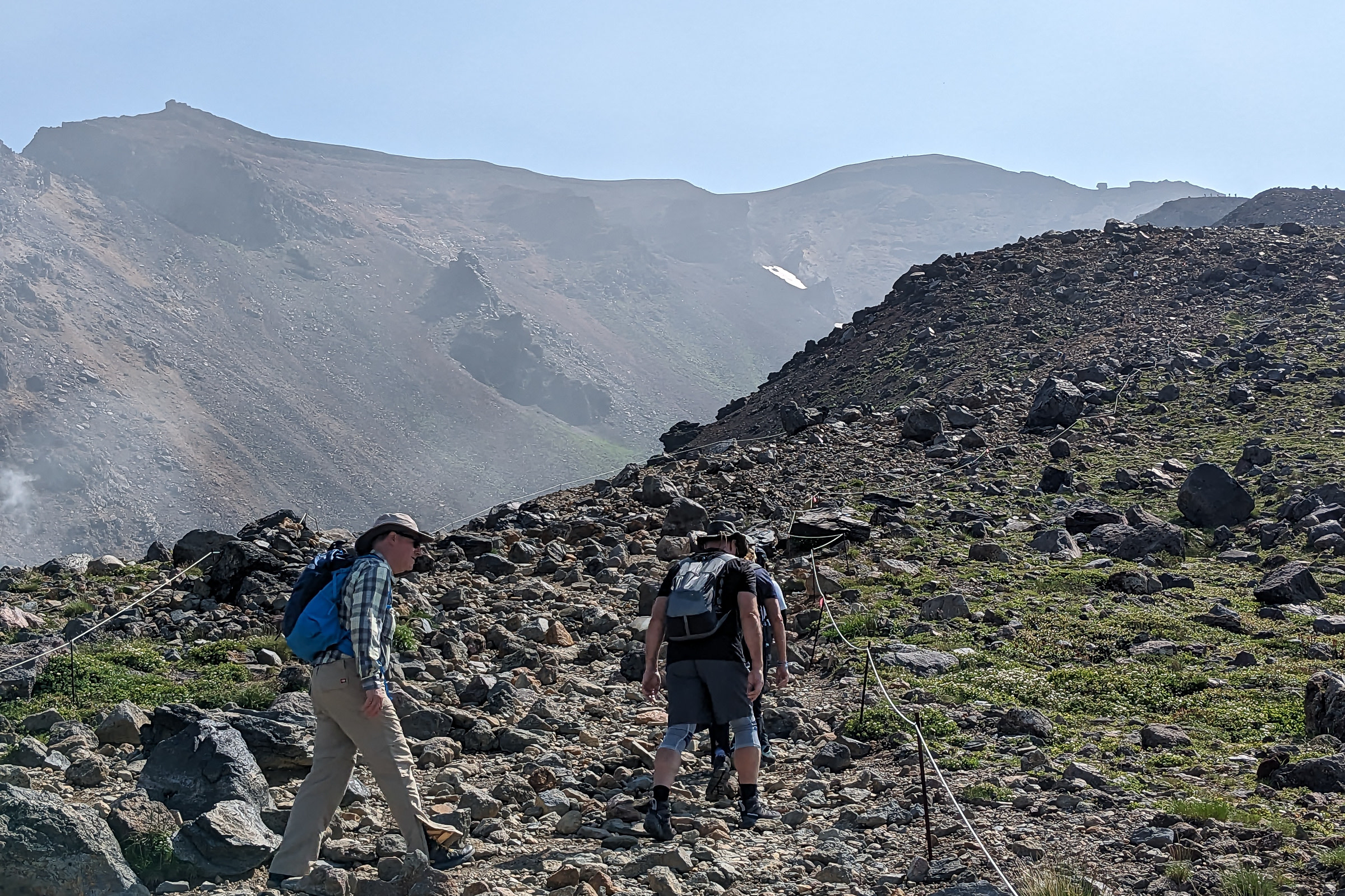 A group of three hikers climb up a rocky path on the way to the summit of Mt. Asahidake. It is a bright sunny day.
