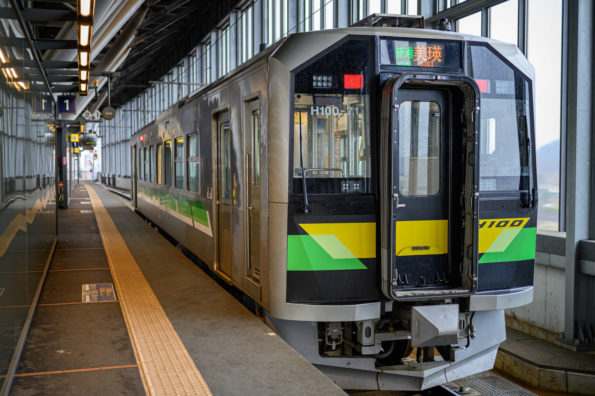 A small, one-carriage train waiting at a platform at Asahikawa Station. The ticker on the front shows it is bound for Biei station.