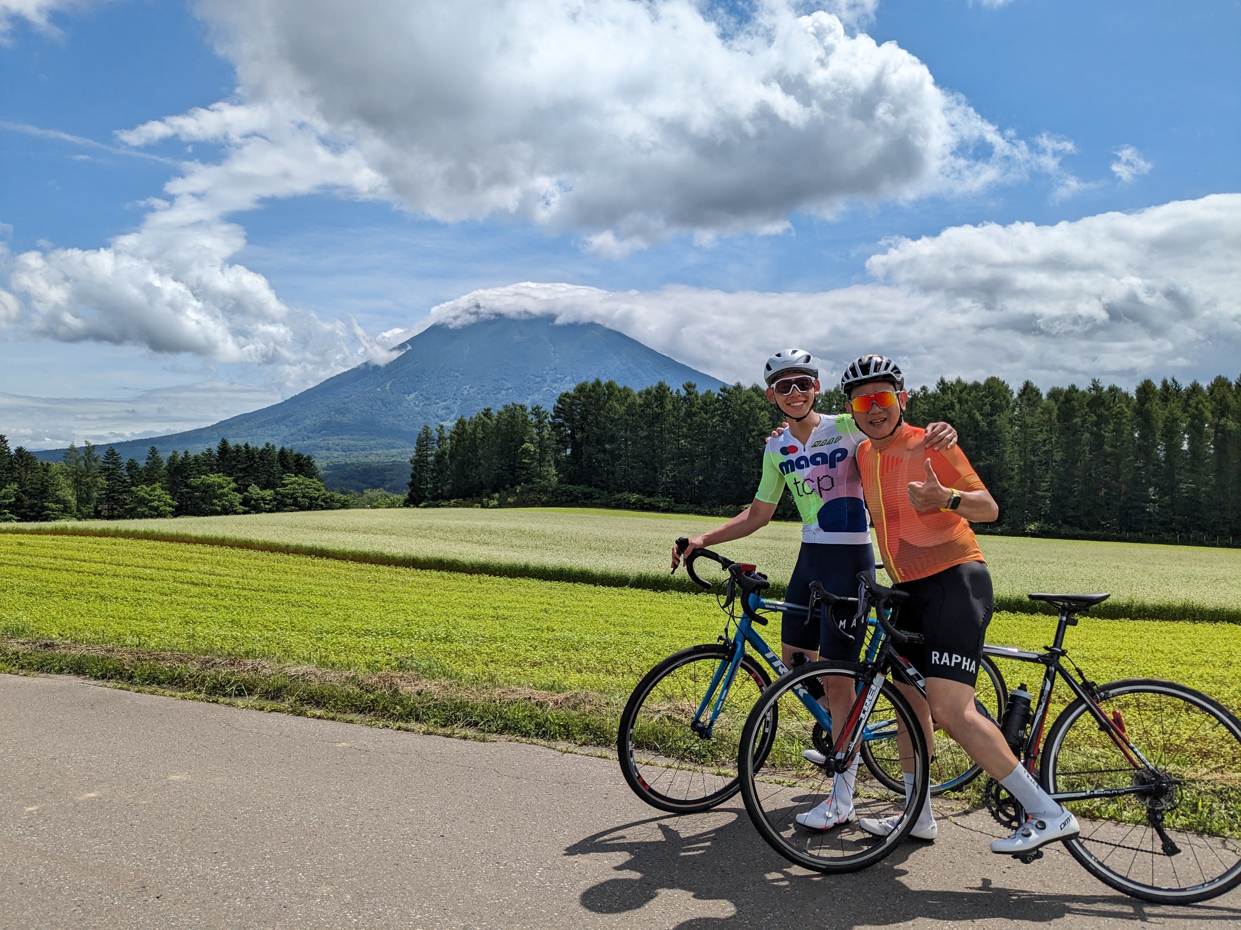 Two cyclists happily pose in front of blooming fields, with Mt. Yotei wreathed in cloud behind them.
