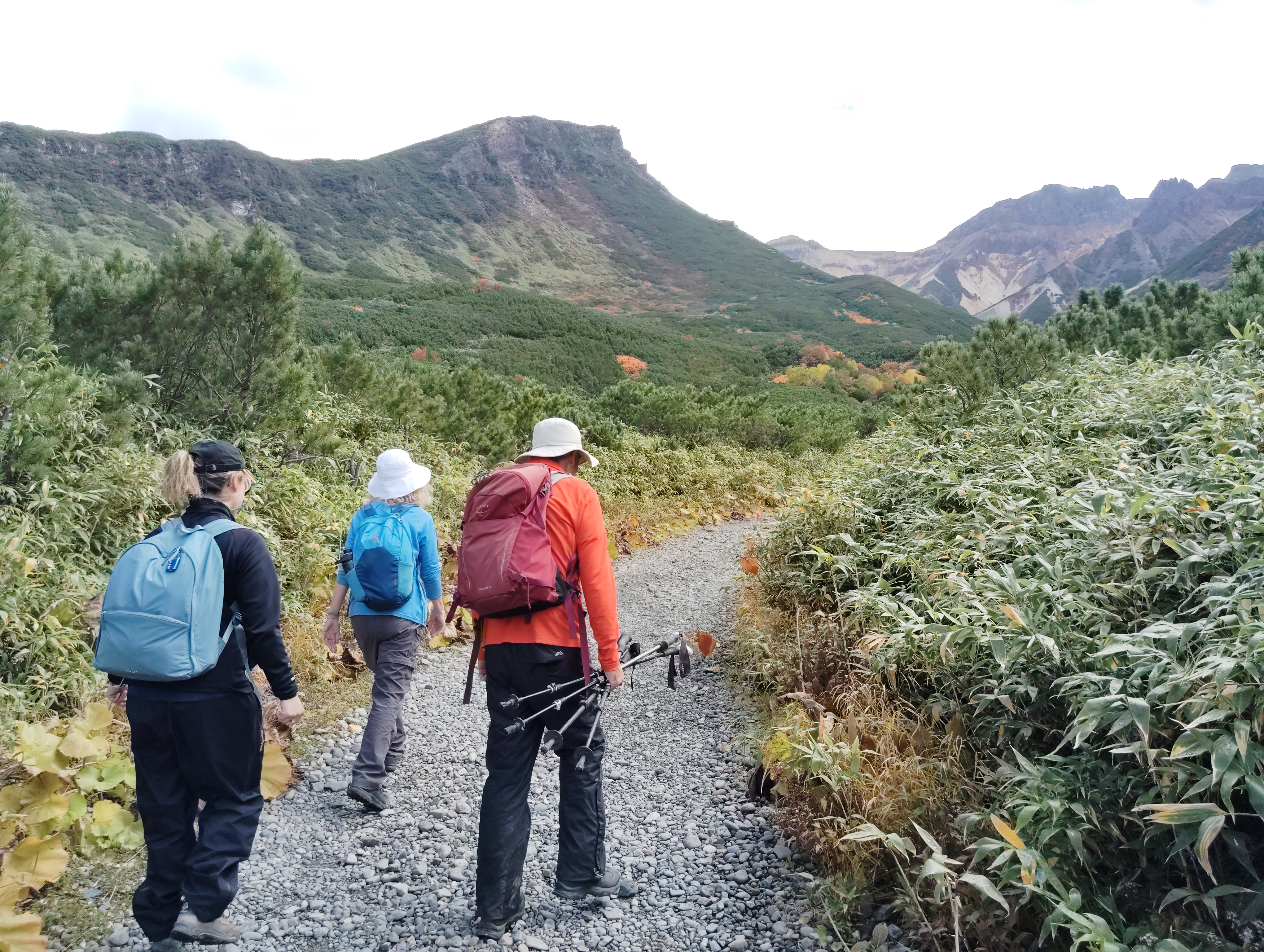 A group of three hikers make their way along a wide gravel path. There is a large mountain pass in the distance and they are heading for it. It is a cloudy day.