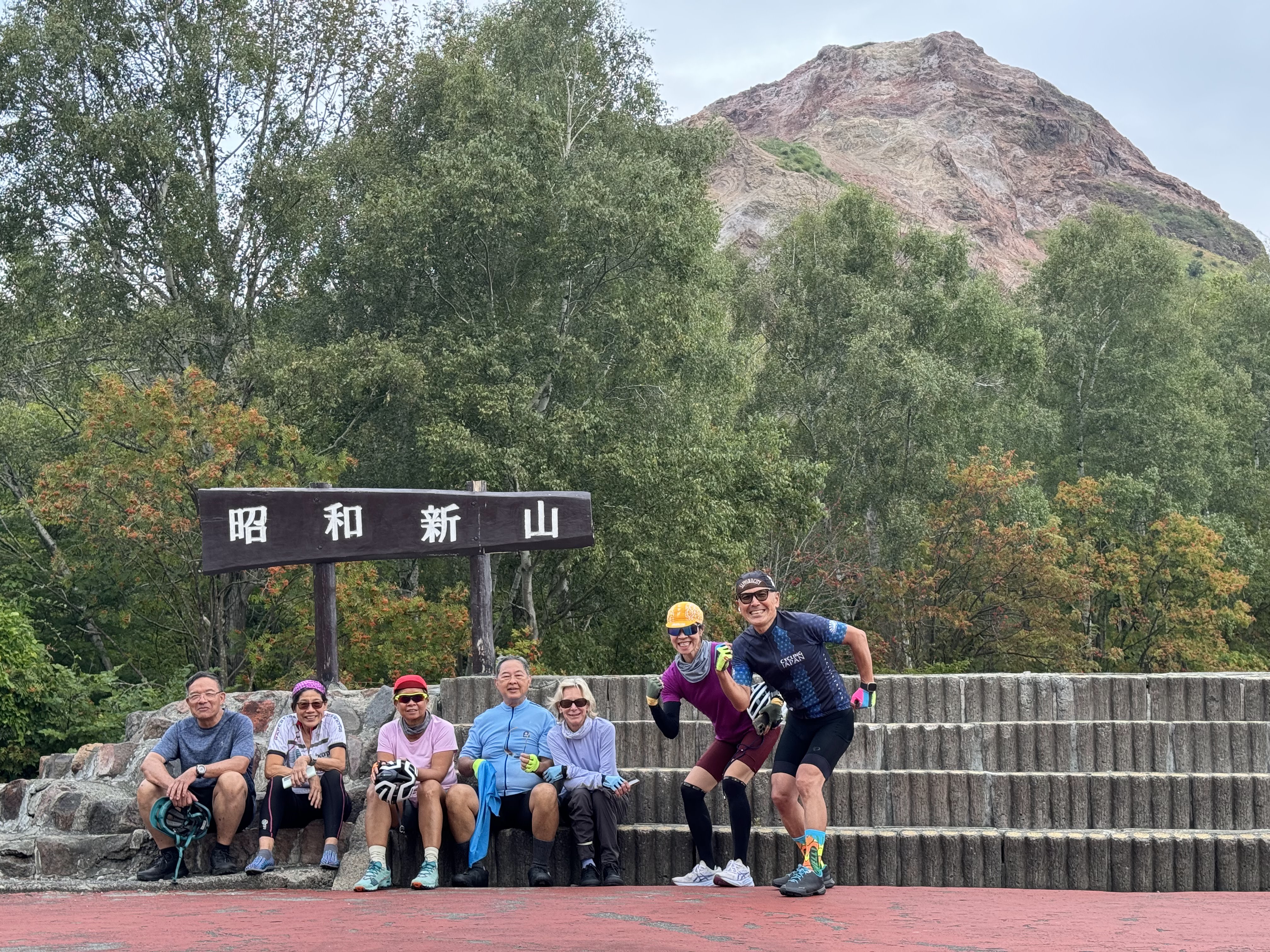 A group of seven cyclists smile at the camera in front of Mt. Showa-Shinzan, an active volcano in Hokkaido. In the background behind them, the volcano's lava dome is visible. 