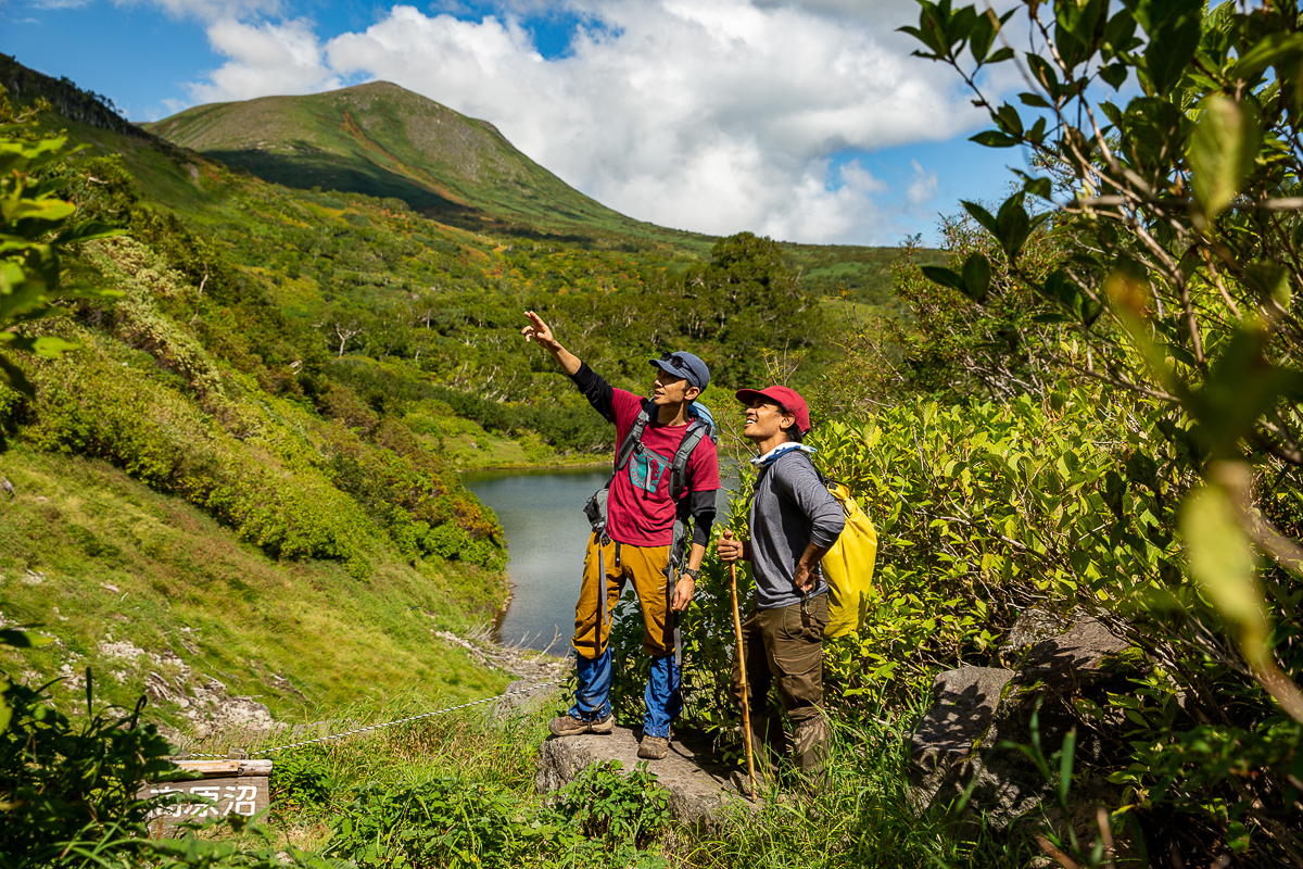 A nature guide points out Brown Bear habitat to a hiker at Kogen Numa in the Daisetsuzan National Park. Behind the pair is a still pond and a large green mountain.