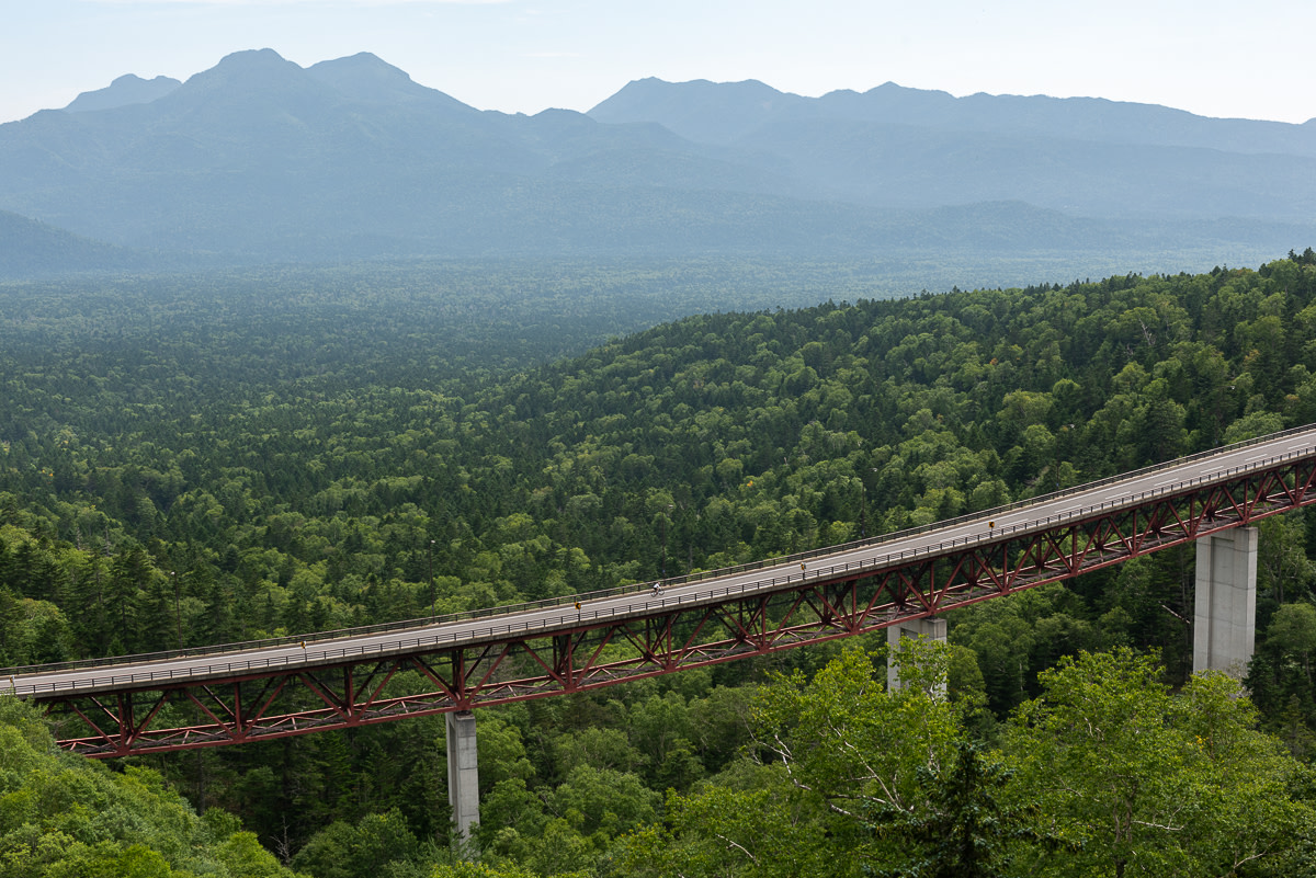 A cyclist riding across a bridge is dwarfed by the scenery of the Daisetsuzan National Park