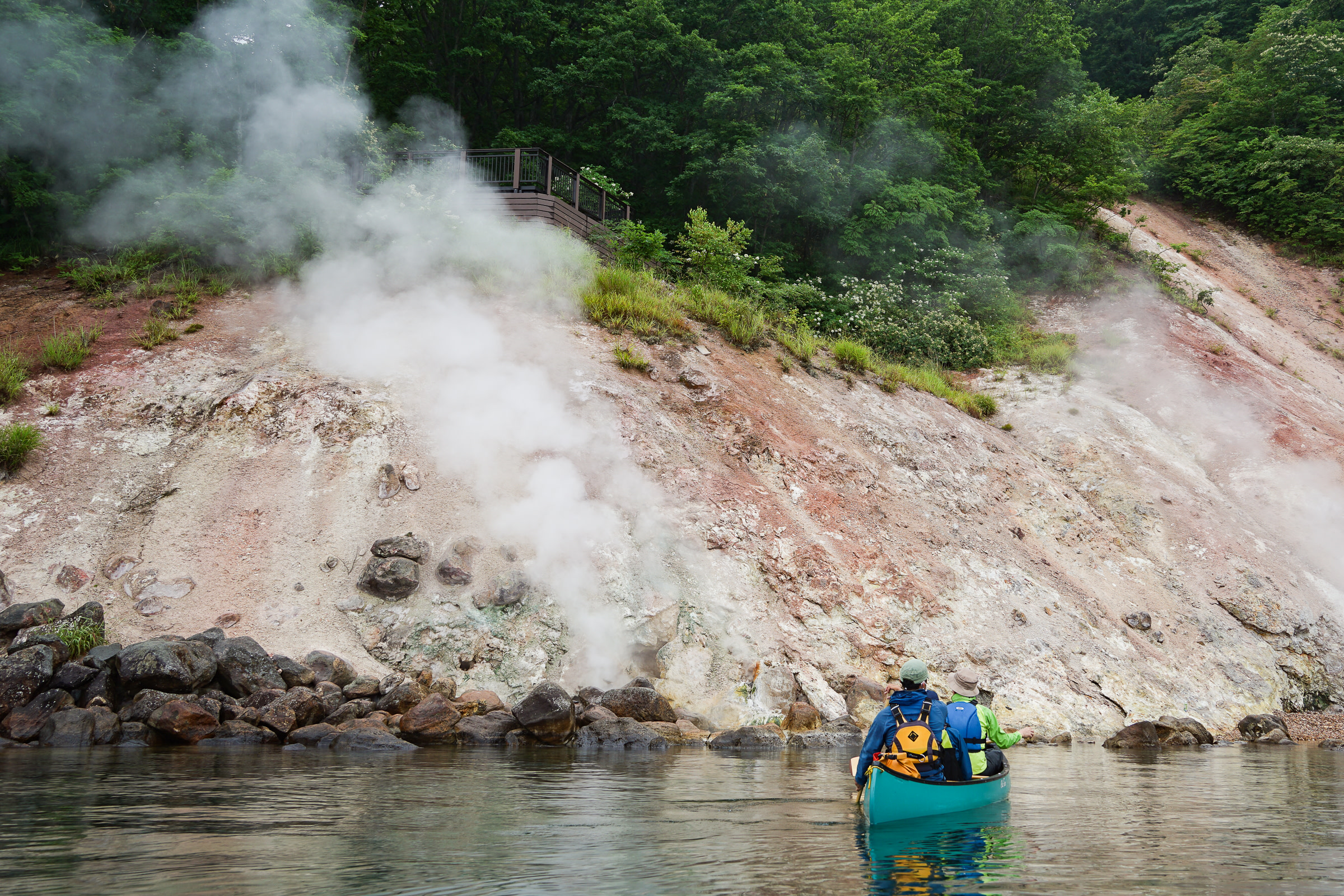 Two canoers approach a steaming hot spring vent on the shore of Lake Kussharo. The vent sits at the bottom of a barren and rugged cliff.