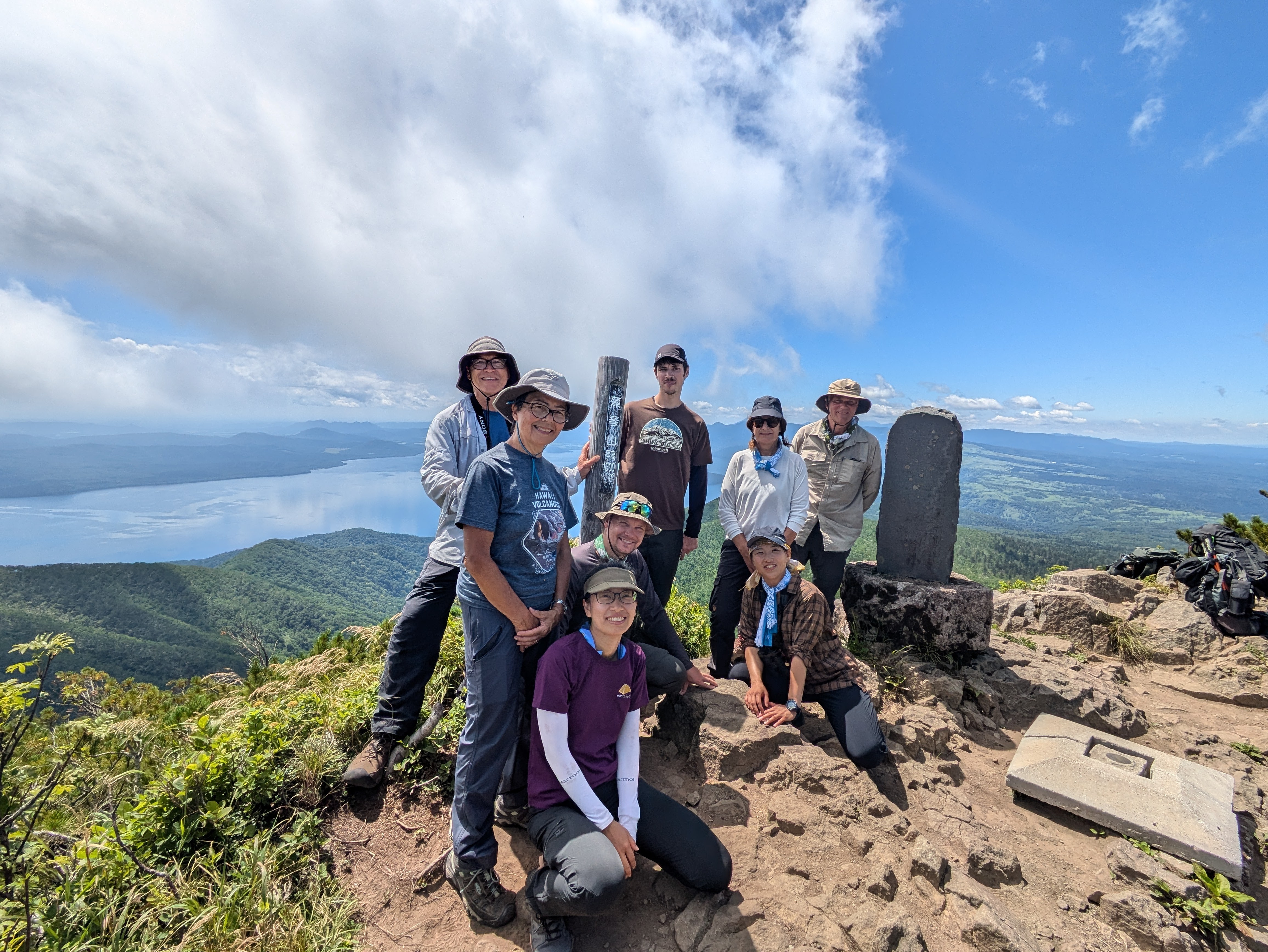 A group of hikers smile at the camera at the summit of Mt. Mokoto in Hokkaido. It is a beautifully clear day and a lake is visible behind them. A pole with Japanese text reads "Mt. Mokoto, 1000m".