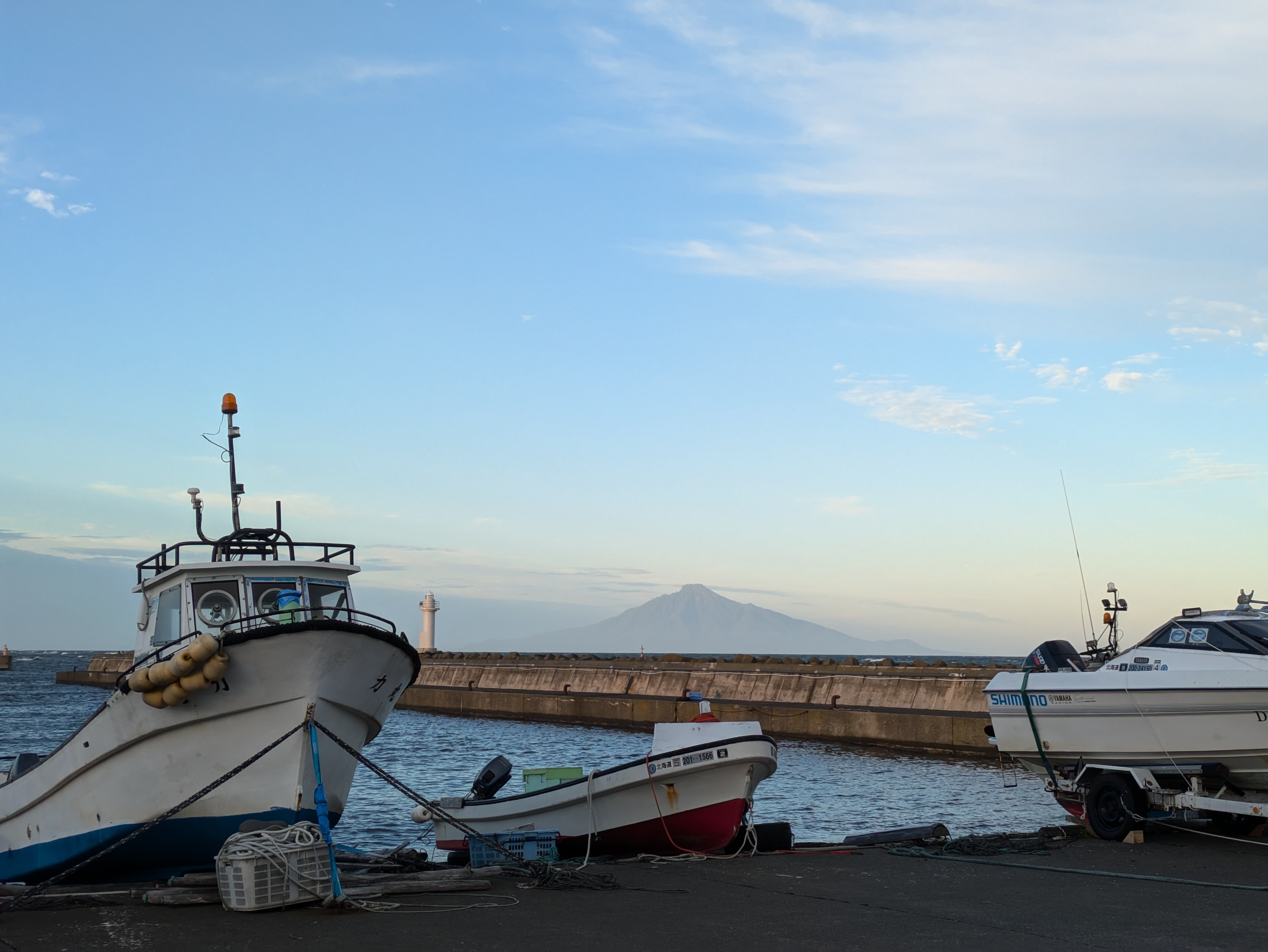 A view of Mt. Rishiri at sundown. It rises over the horizon, with boats moored on the coast in the foreground.