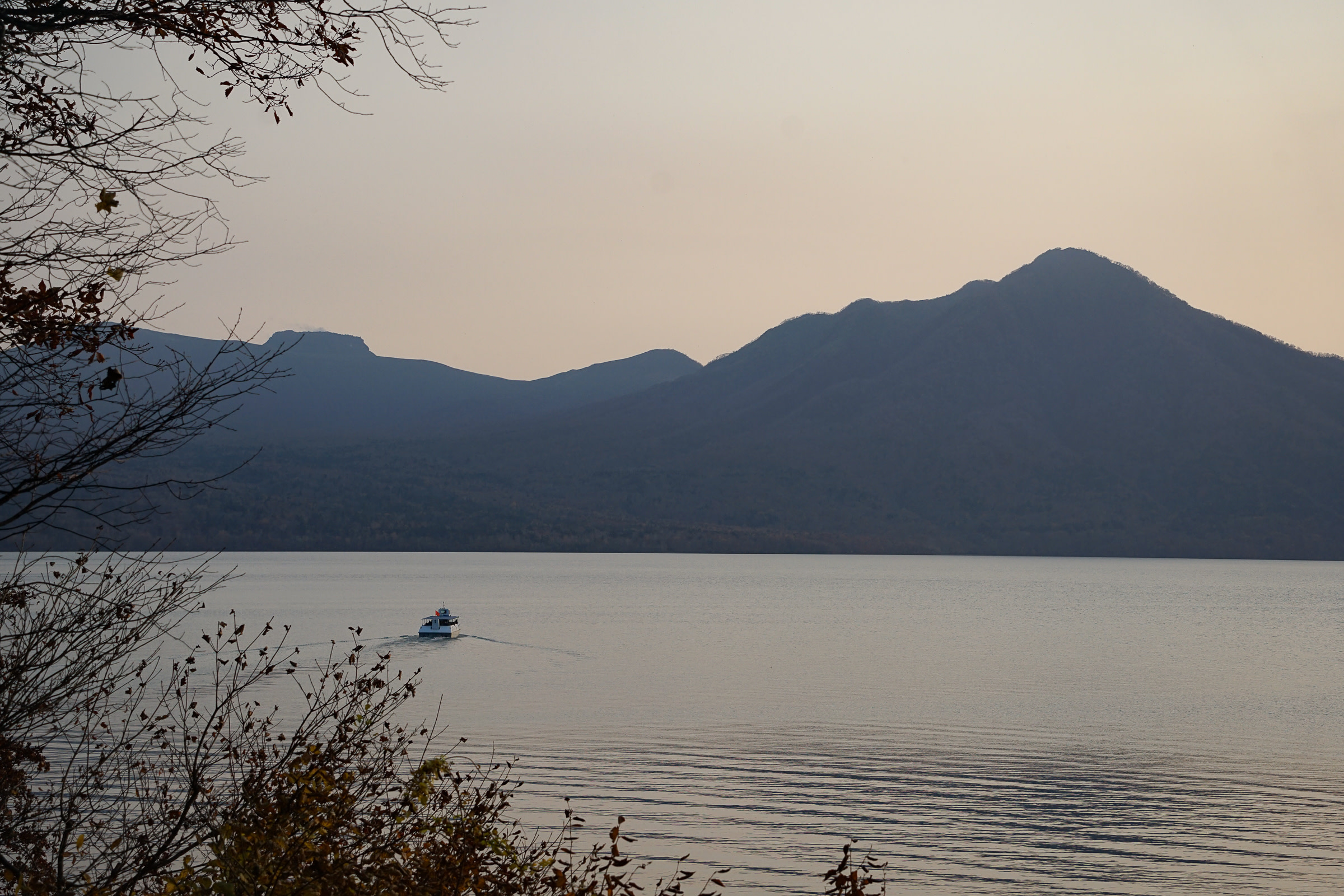 A ferry in Lake Shikotsu at dusk goes in the direction of dark mountains in the distance.